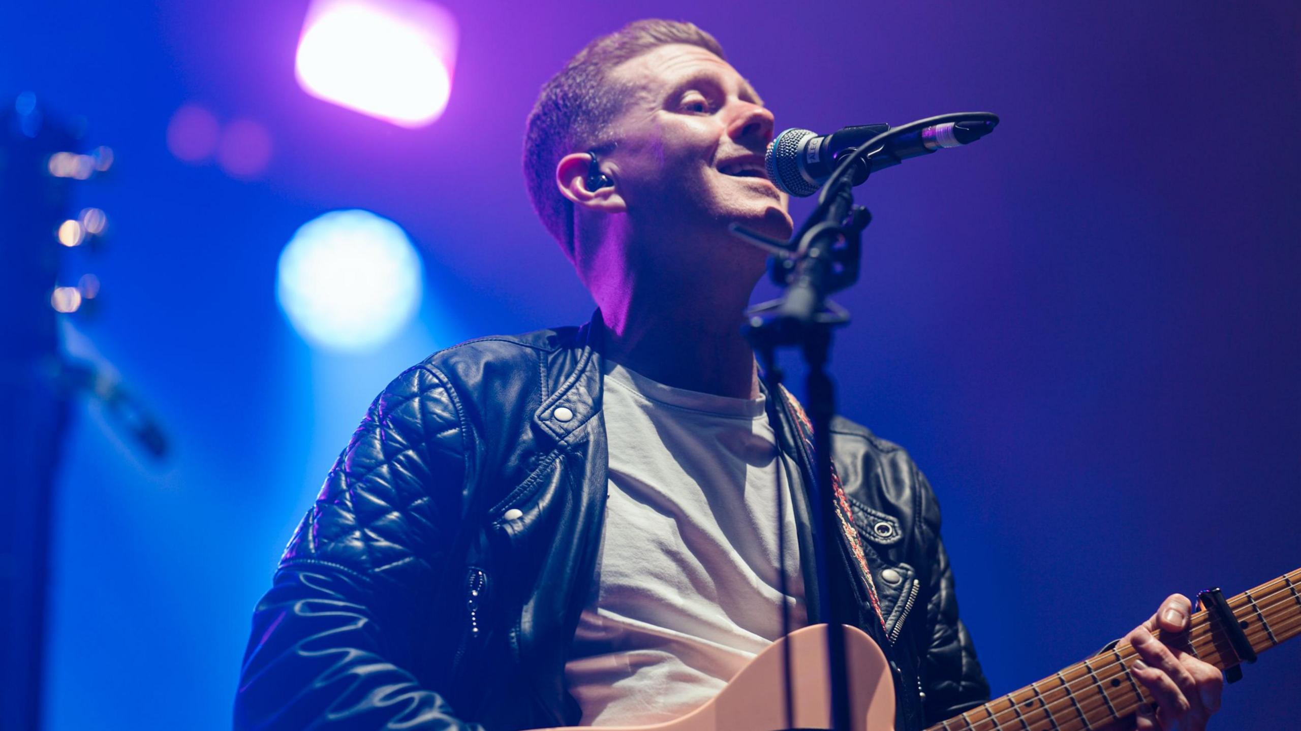 Man singing into a mic and playing guitar. He is wearing a shit T-shirt and black leather jacket and there are blue lights behind him