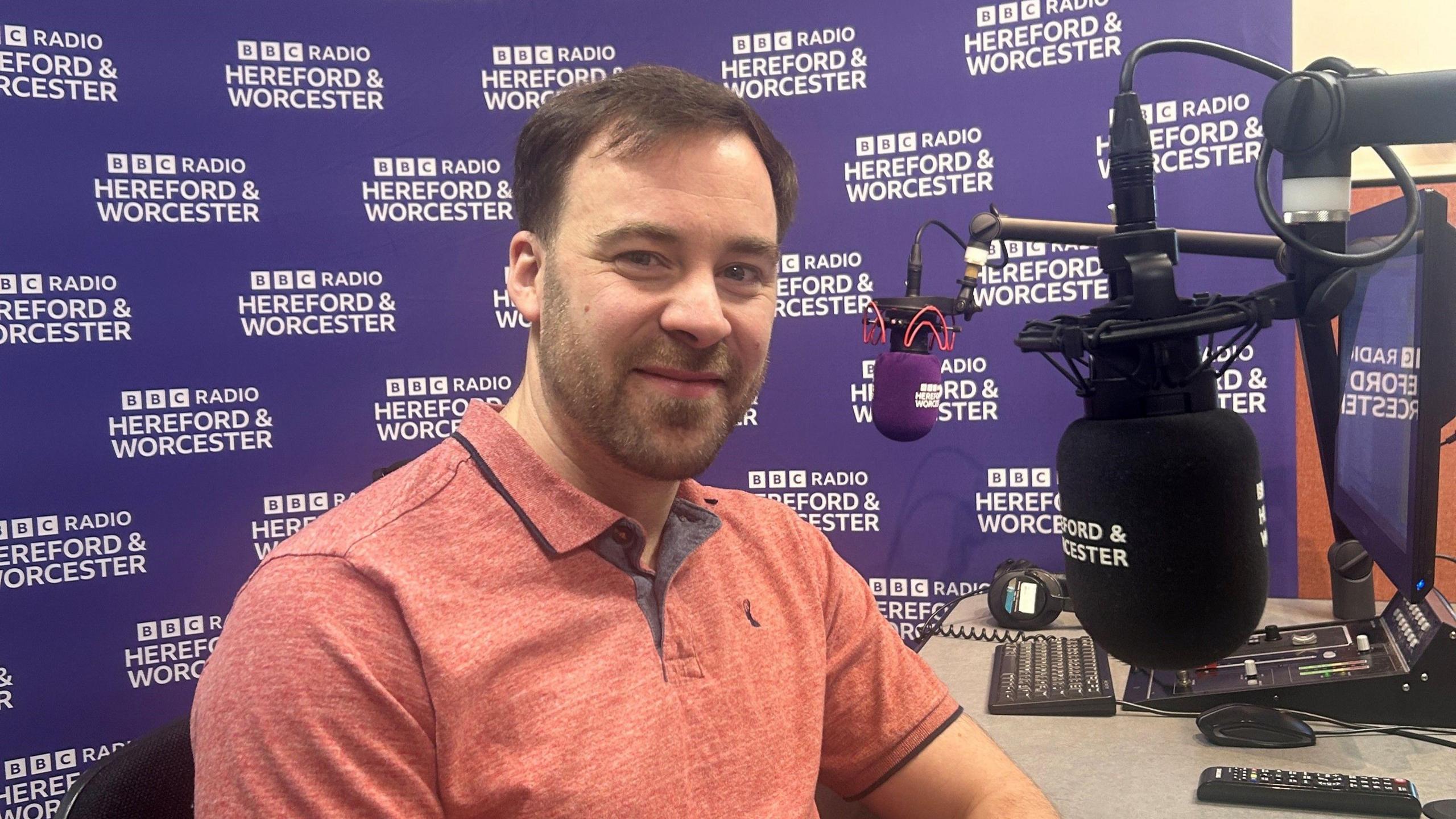 Mr Perrett is sitting at a desk with a black radio microphone in front of him; He has brown hair and brown facial hair, and is wearing a salmon pink T-shirt. In the background is a purple bbc hereford and worcester banner