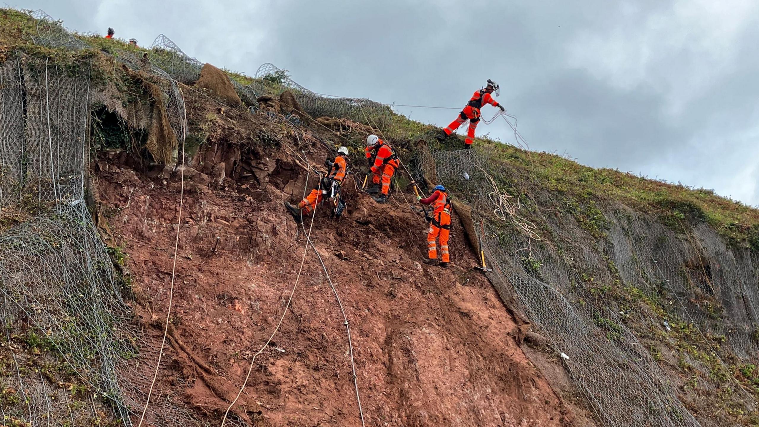Network Rail workers on slopes using harnesses. The five workers are wearing orange uniforms and work hard hats and are attached to the harnesses. There is also netting on the slopes which are brown with grass on the top.