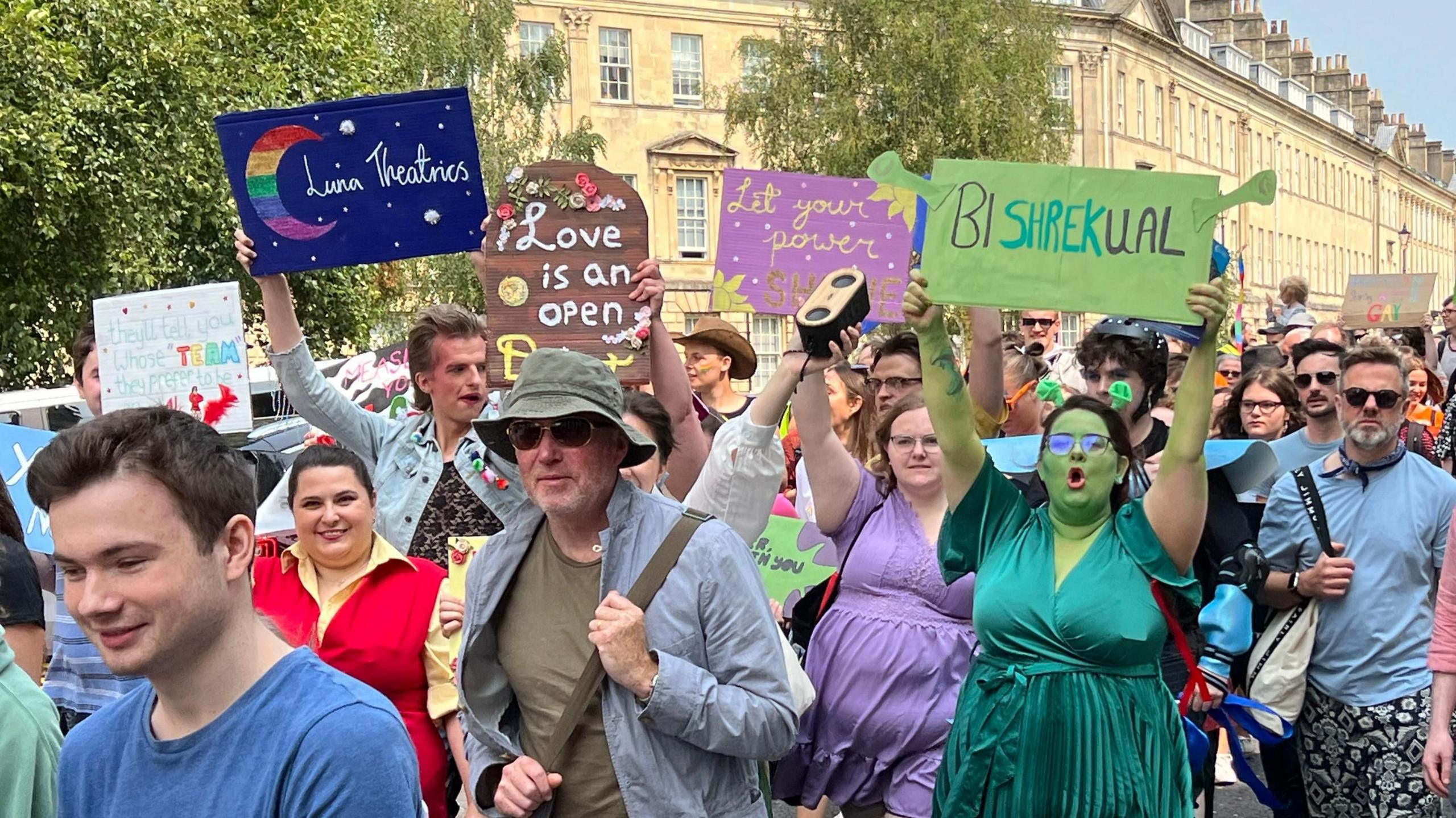 A large group of people holding signs as they march through Bath city centre. One of the signs says 'Love is an open door', another, which is held by a woman dressed as Princess Fiona from the film Shrek, says 'Bi-Shrek-Ual'.