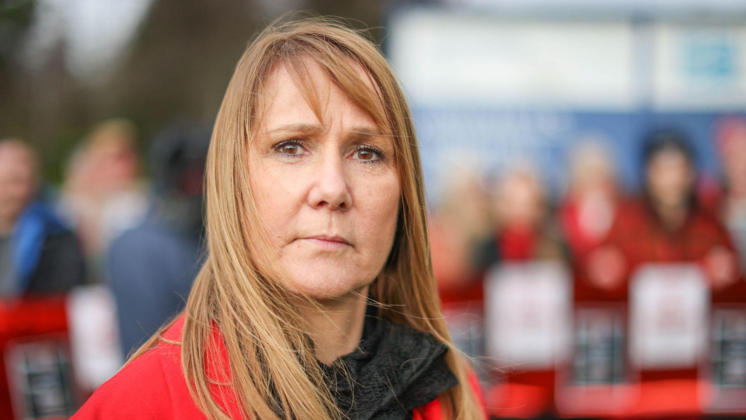 Jules Rose looks into the camera - she has long brown/blonde hair down to her shoulders, a concerned expression on her face and a red jacket on.  In the background other people can be seen, likely fellow protestors against the surgeon, but the image is blurry so their faces can't be seen.