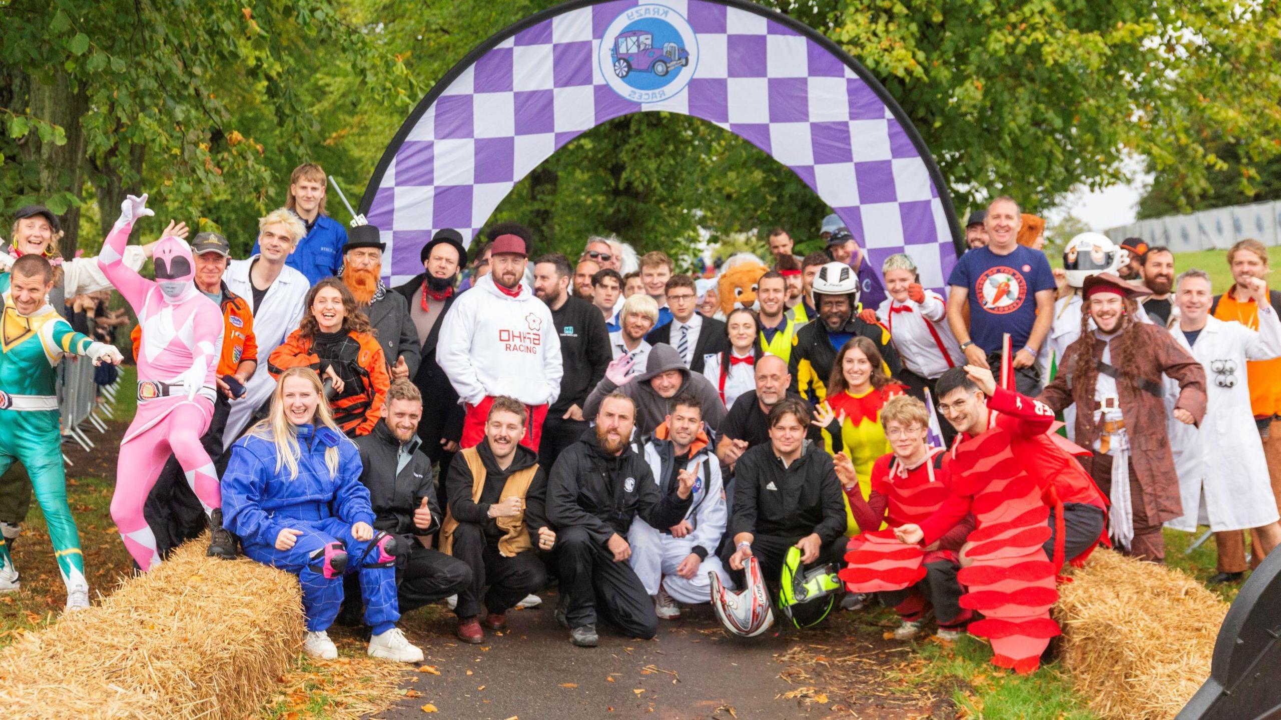 A large group of people, many of them in fancy dress costumes, stand under a finish line hoop which has a purple check pattern. A row of straw bales is seen on each side of a tarmac path leading up to where the people are standing. The event is a Krazy Races meeting in Victoria Park in Bristol.