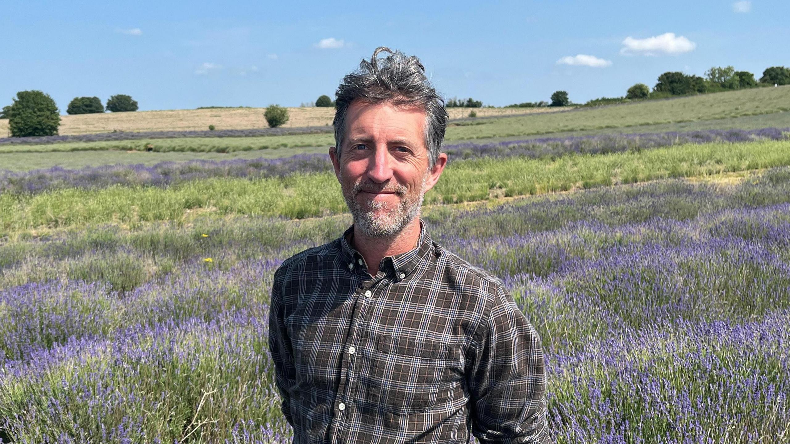 Tim Hunter with a checked shirt standing in a lavender field. He has brown wavy hair and is smiling as he looks into the camera.