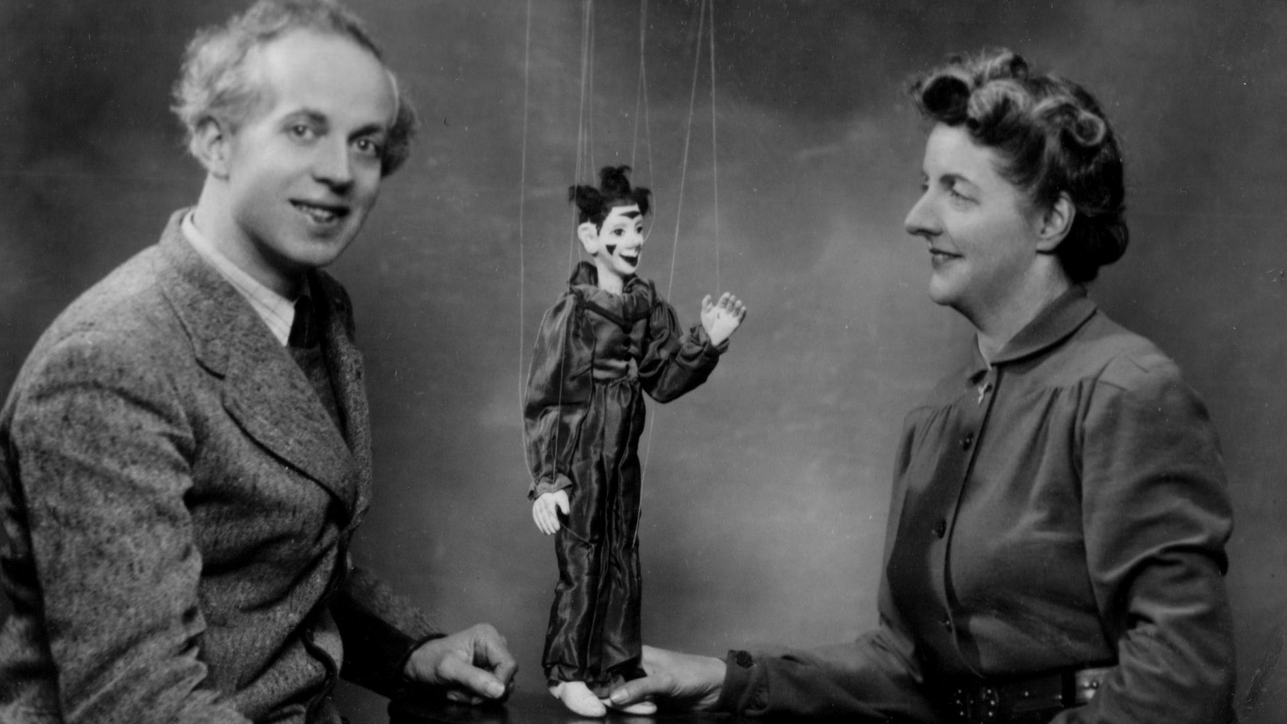 A black and white photograph showing a man and a woman sitting either side of an old-fashioned marionette puppet with strings attached.