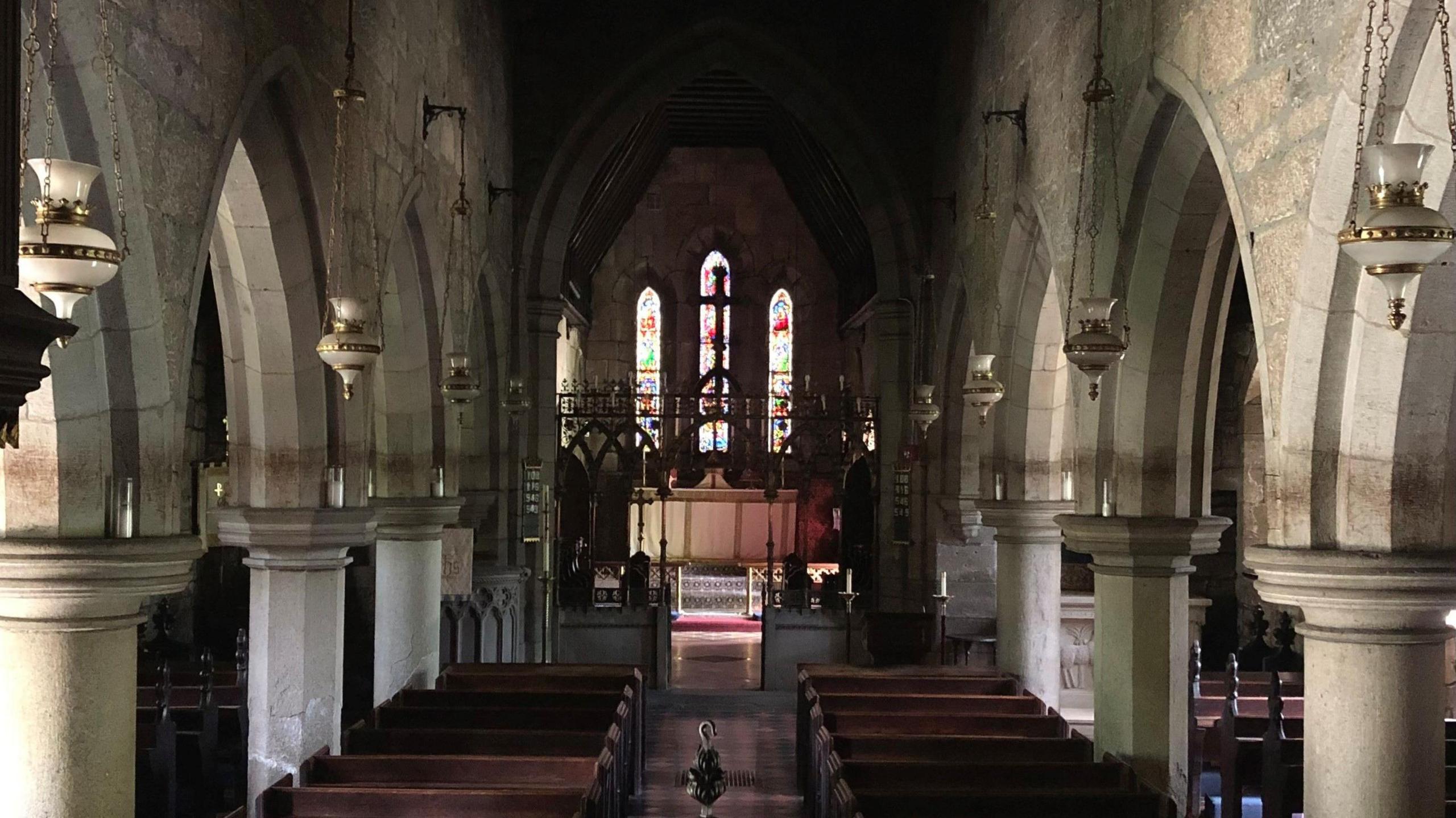 The inside of a church shows wooden pews and a tiled floor. There are stone arches to the sides and a stained glass window at the end