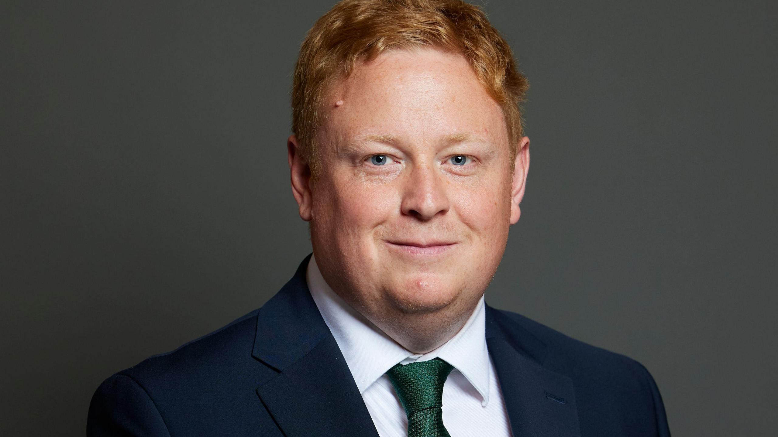 A man with ginger hair wearing a navy suit jacket, white shirt and green tie smiles at the camera