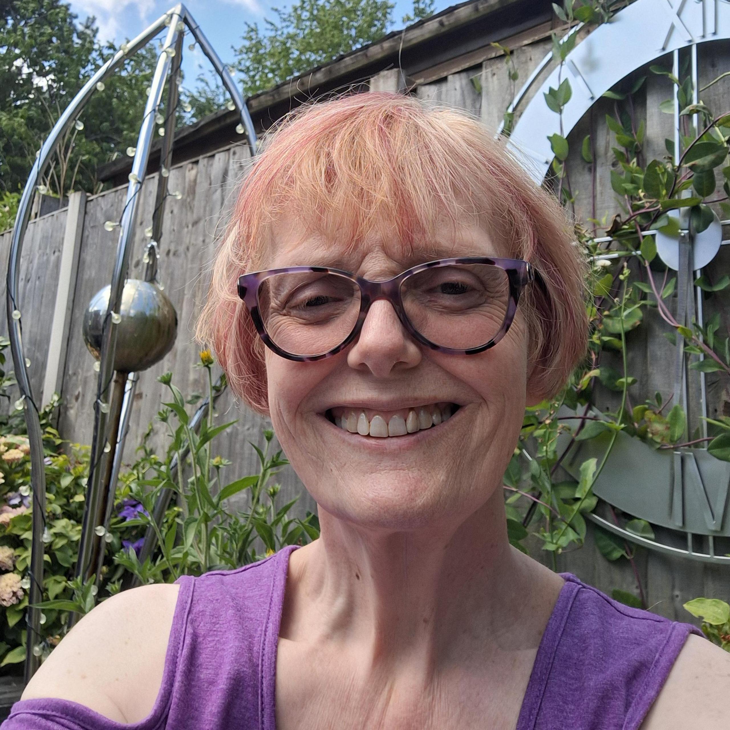 Jo Bain wearing glasses and a sleeveless purple top in the foreground, facing the camera in a garden setting. Behind her are flowering plants, climbing greenery, a wooden fence and a large decorative outdoor clock mounted on the fence. A metal garden ornament and blue sky with scattered clouds are visible in the background.