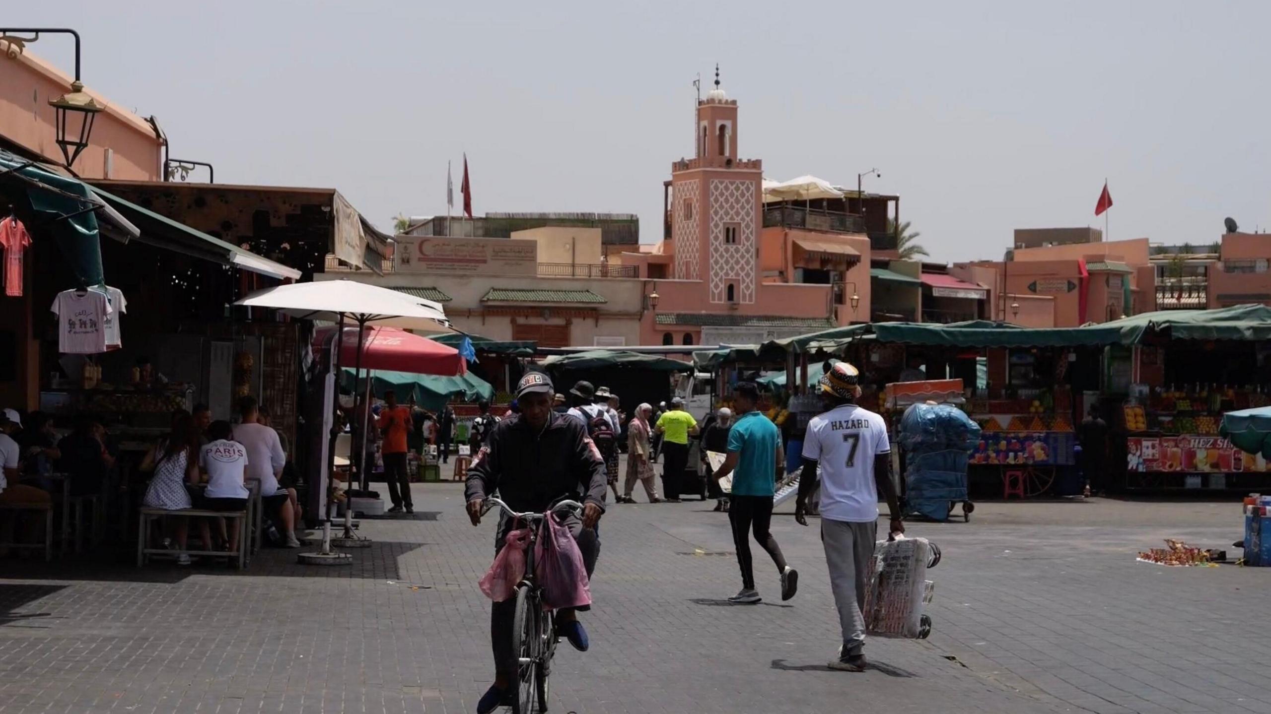 A busy market in Marrakesh filled with both tourists and locals. There is a mana on a bike, people sitting down and dining, and people walking around with shopping bags.