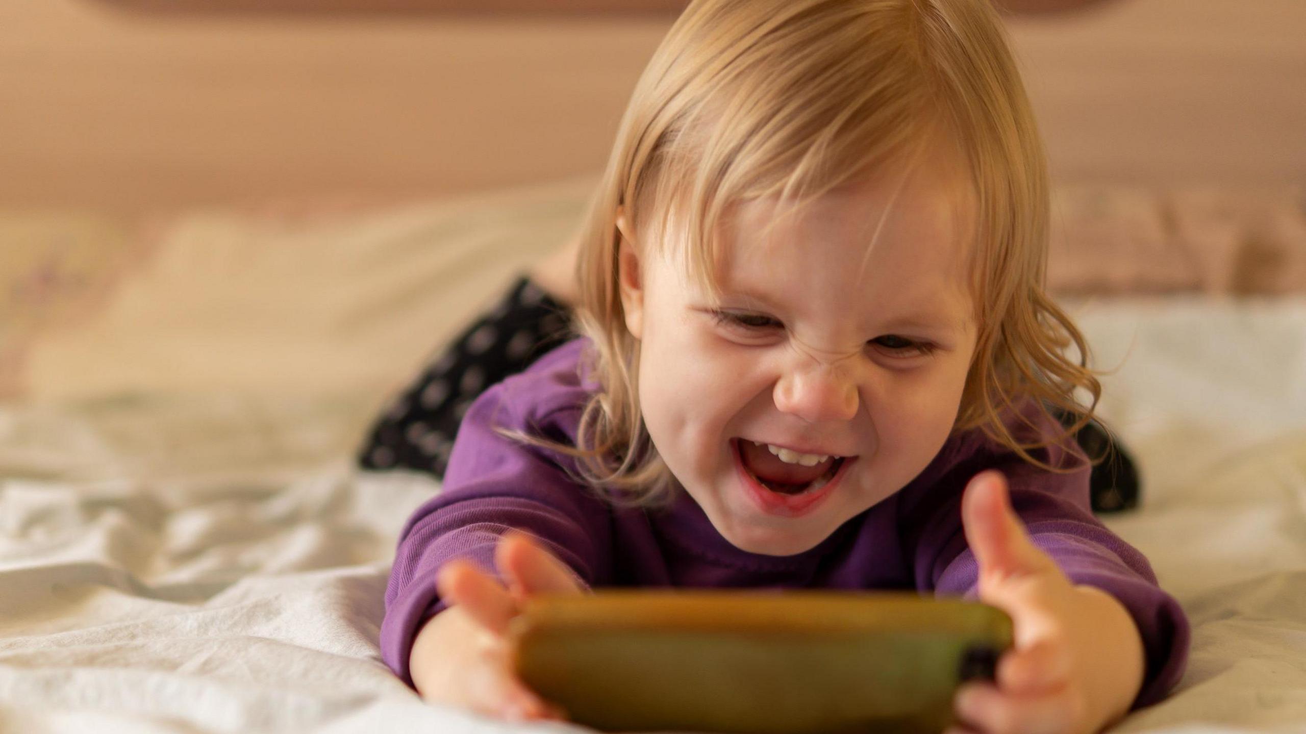 Girl with blonde hair aged about three is lying on her stomach on a bed with white bedsheets. She is holding a phone in her two hands and is smiling with her nose scrunched. She looks happy.