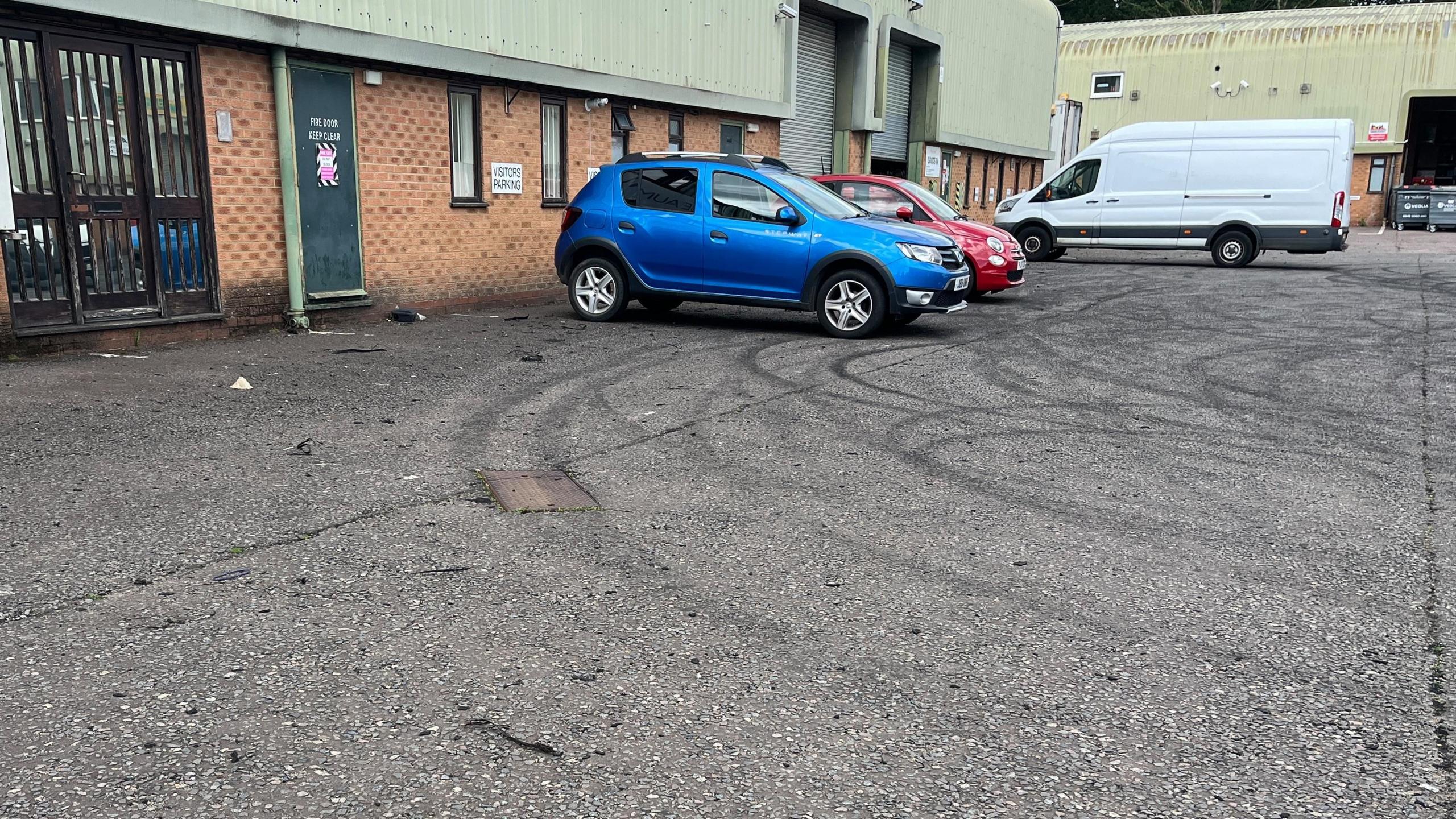 Burning rubber tyre marks on the ground in an industrial estate. The circular patterns are close to the edge of the buildings. There are two cars and a van in the background.