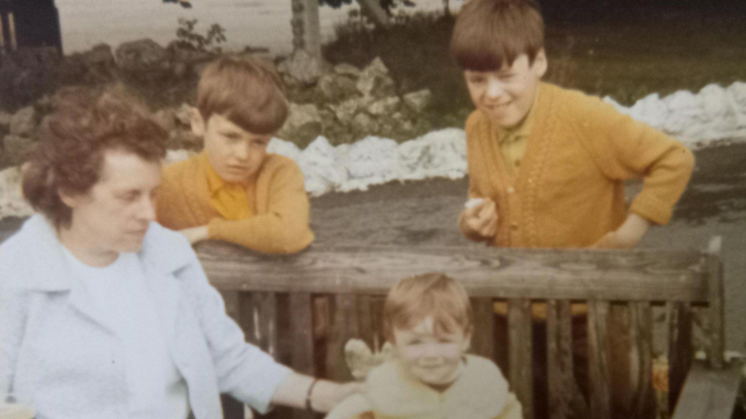 A vintage photo of three young boys and a woman. They are sitting on and standing behind a bench.