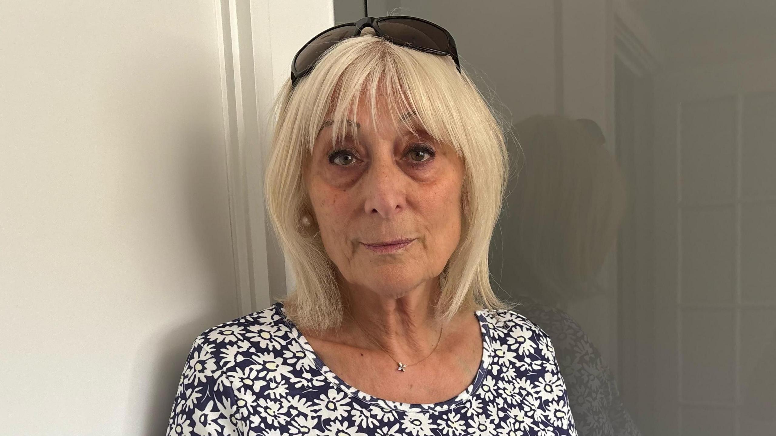 Carol Ferrary is wearing a black and white flowery top. She has blonde hair and sunglasses on her head, and has a serious expression on her face.