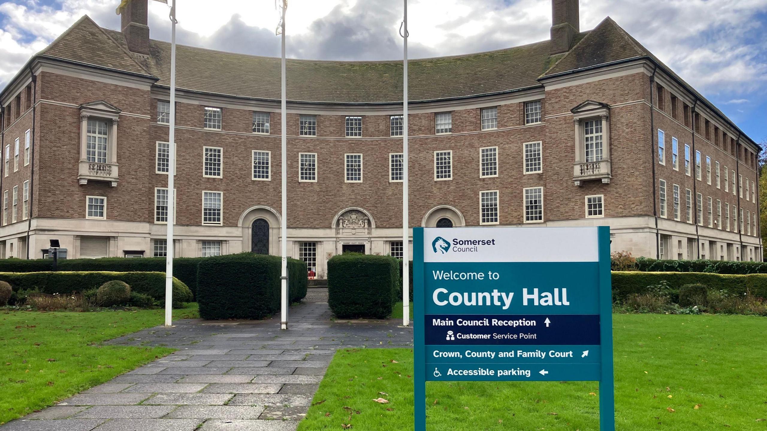 Photo of the County Hall building in Taunton, including a Somerset Council sign and flagpoles  in the foreground. The brick building has a curved face and is four stories tall with large windows and ornate stonework around the lower floor. 