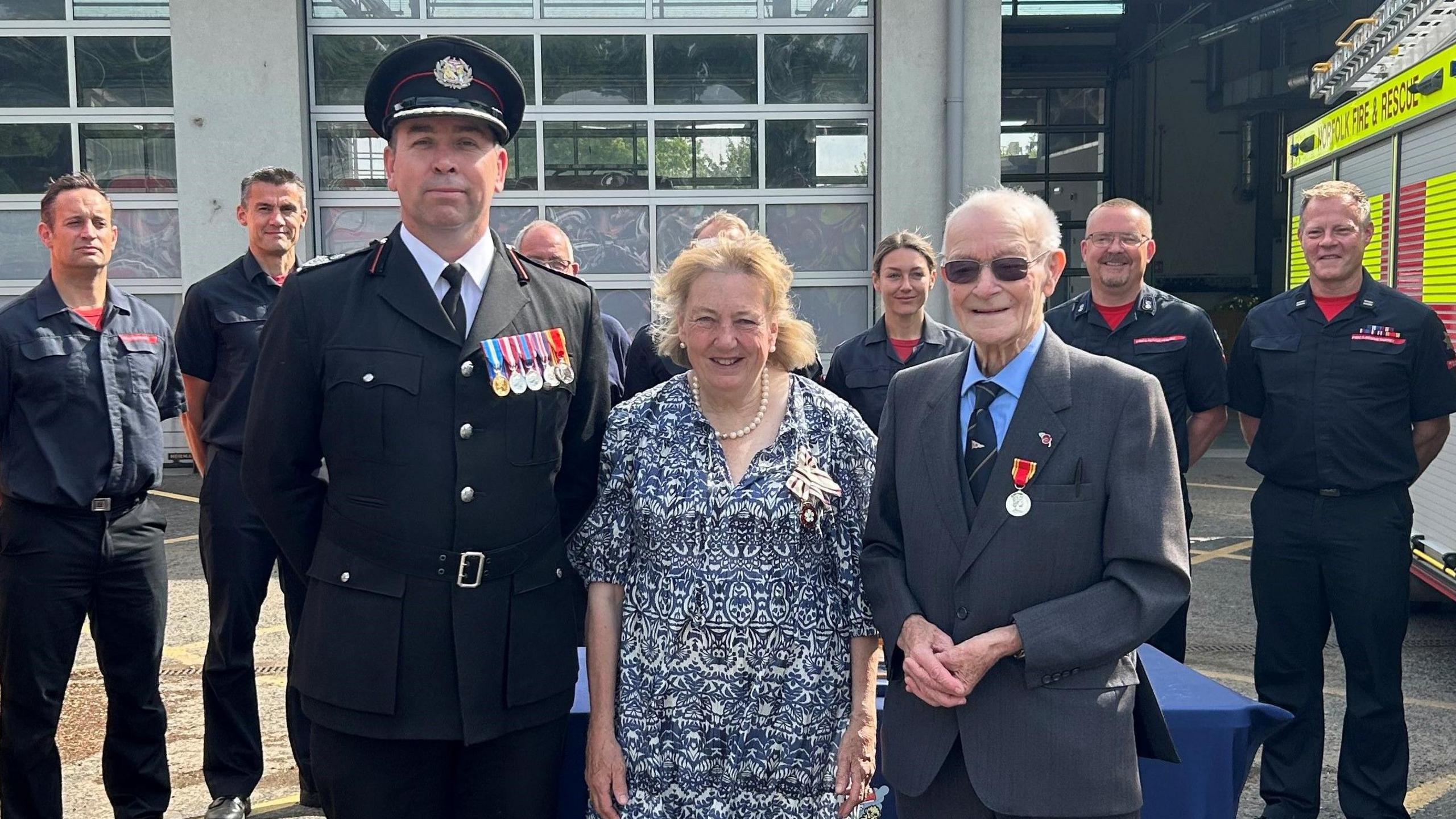 Three people are standing in the forefront of the image. Two men are on the ends wearing suits with medals on their jacket, and a woman is standing in the middle wearing a blue dress. People in uniform are standing in the background.