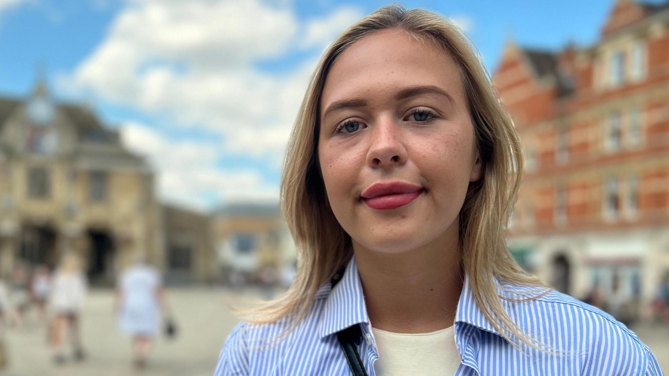 Daisy Blakemore-Creedon has blonde hair and is standing outside in Peterborough city centre in front of the Guildhall building. She is wearing a butter yellow coloured T-shirt with a white and blue striped shirt over the top. She is looking directly at the camera and the sky is blue in the background behind her.