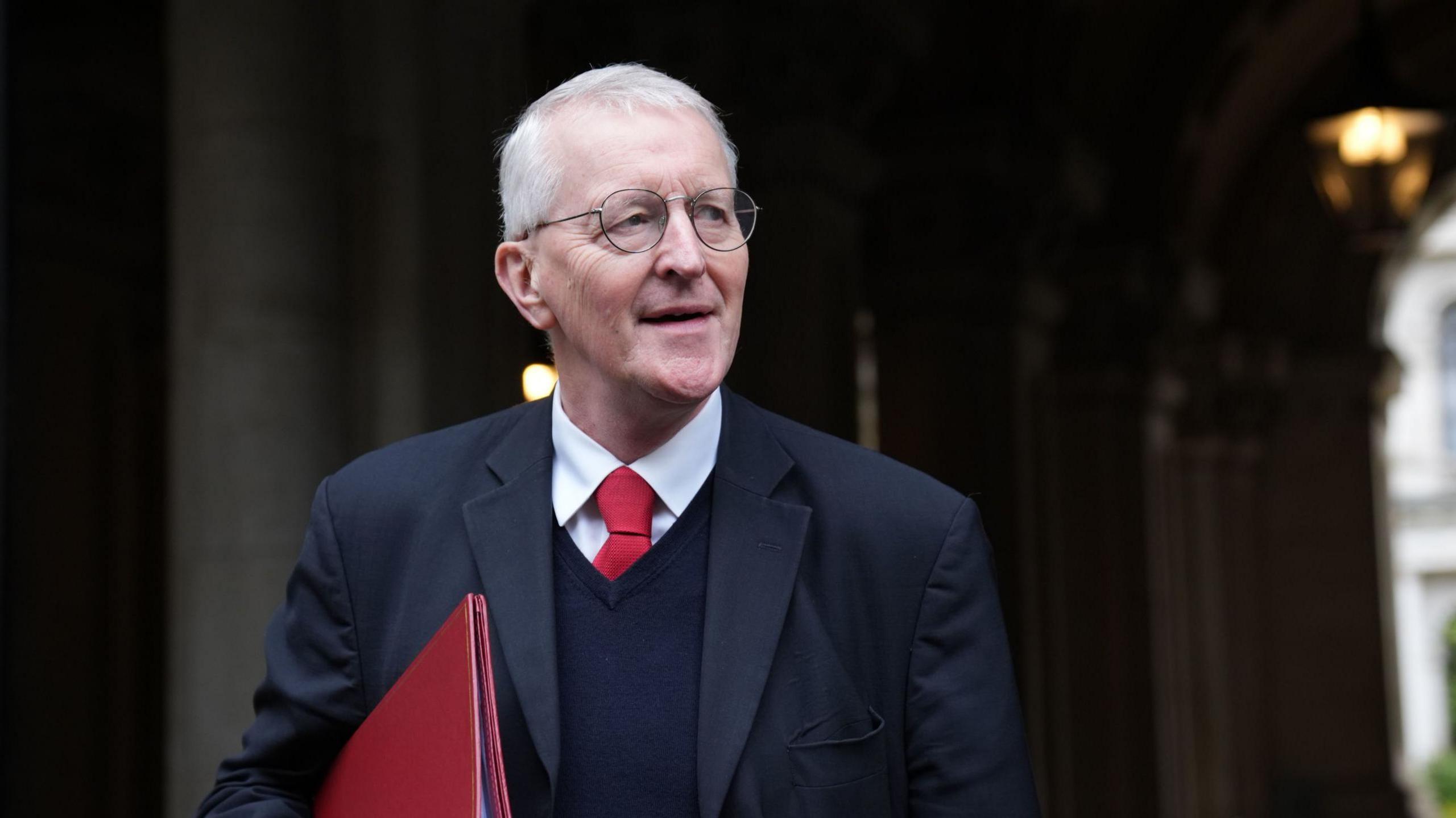 Hilary Benn stands in holding a red folder. He has glasses and grey hair. He is wearing a black suit with a red tie.