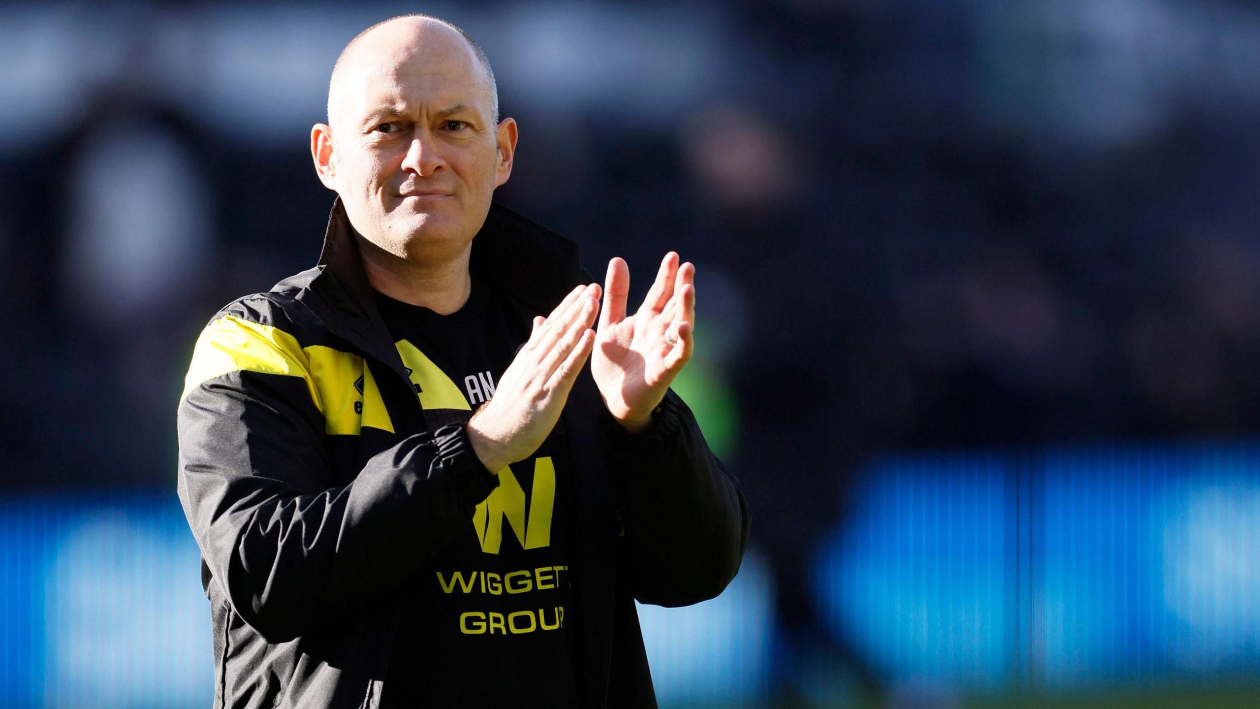 Millwall head coach Alex Neil walks across the pitch at the end of a game clapping the fans in his black and yellow training jacket