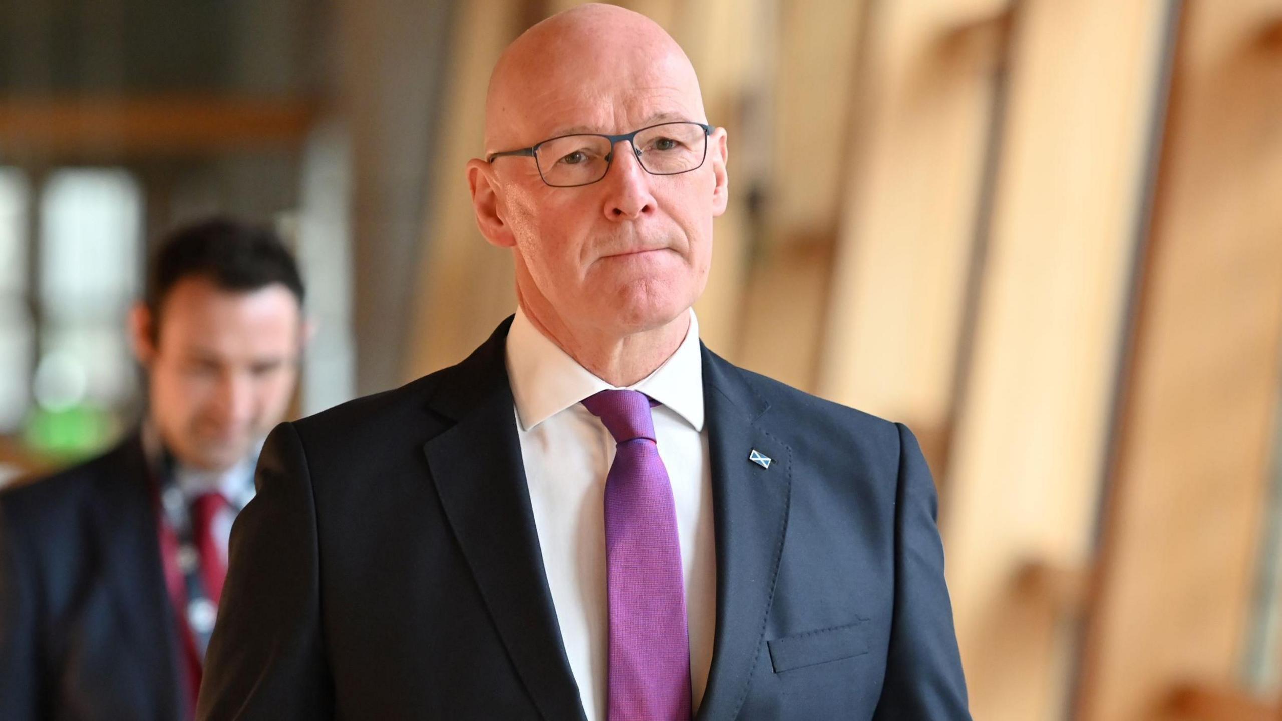 Scotland's first minister, John Swinney, walks through the Scottish Parliament, wearing a dark suit, white shirt and a purple tie.