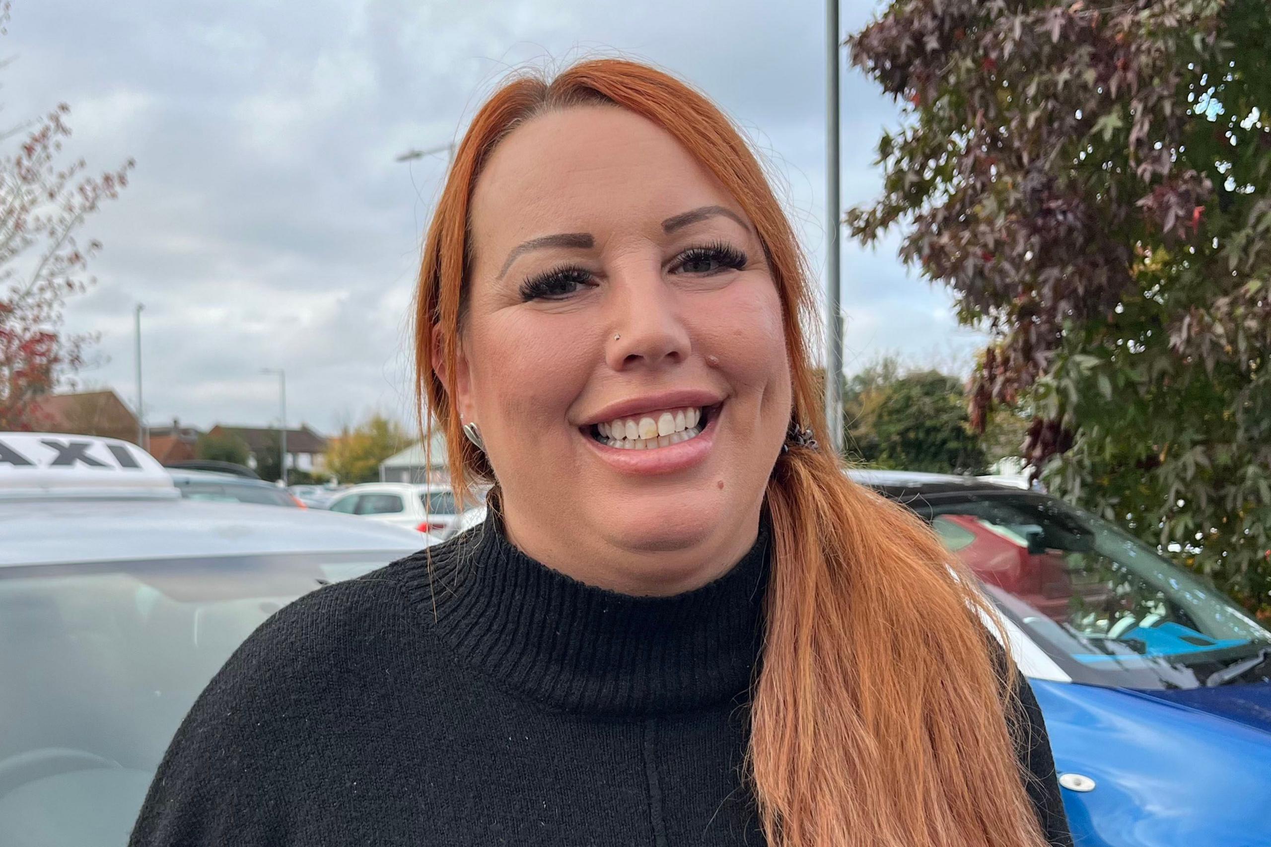 Taxi driver Jenny Wilson smiling at the camera, with a car park in the background. She has long auburn hair tied back in a pony tail and is wearing a black jumper.