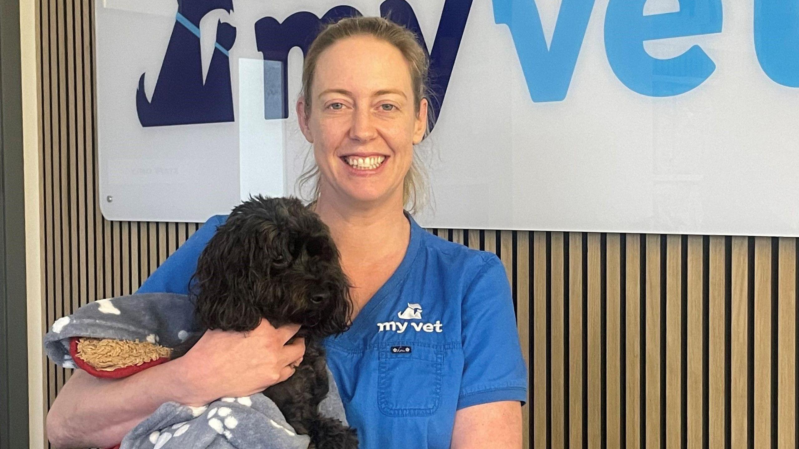 A vet is wearing a blue uniform and holding a small black dog. The vet's hair is fair and pulled back into a pony tail. The dog is wrapped in a blanket.