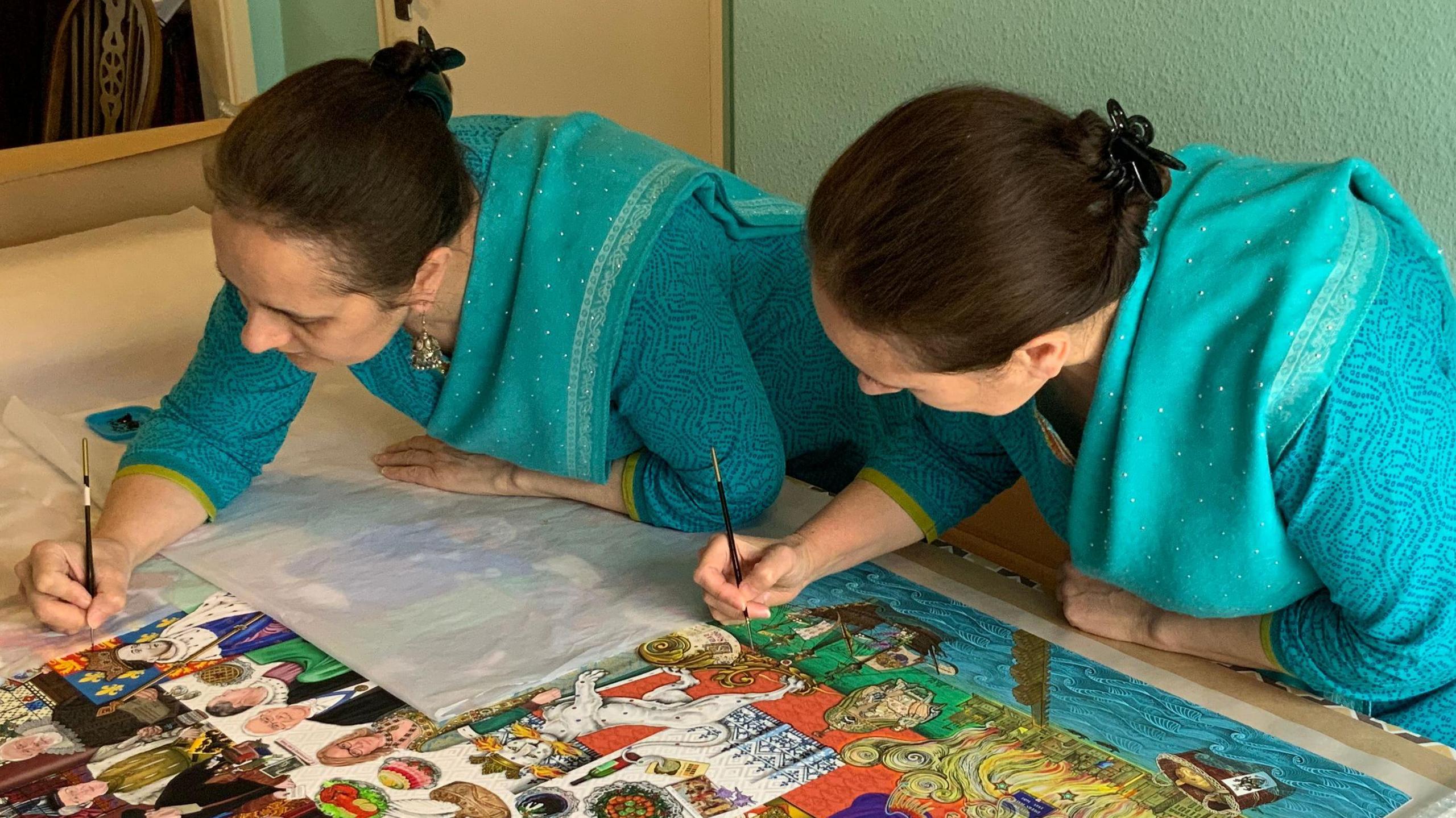 The sisters, dressed in turquoise saris, lean over the same large painting on a table and using fine brushes, add to the details.
