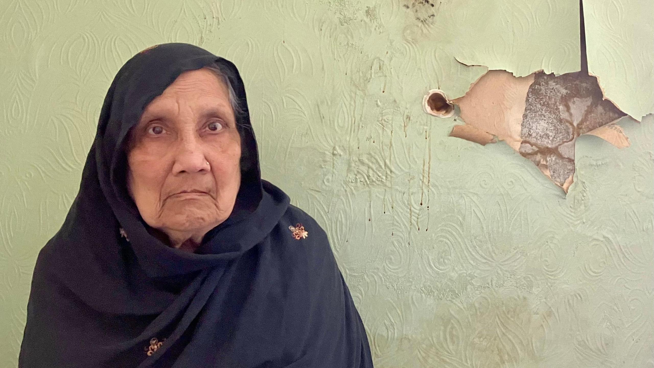 Tormooja Khatun with her head covered, standing in front of peeling wallpaper caused by damp in her house