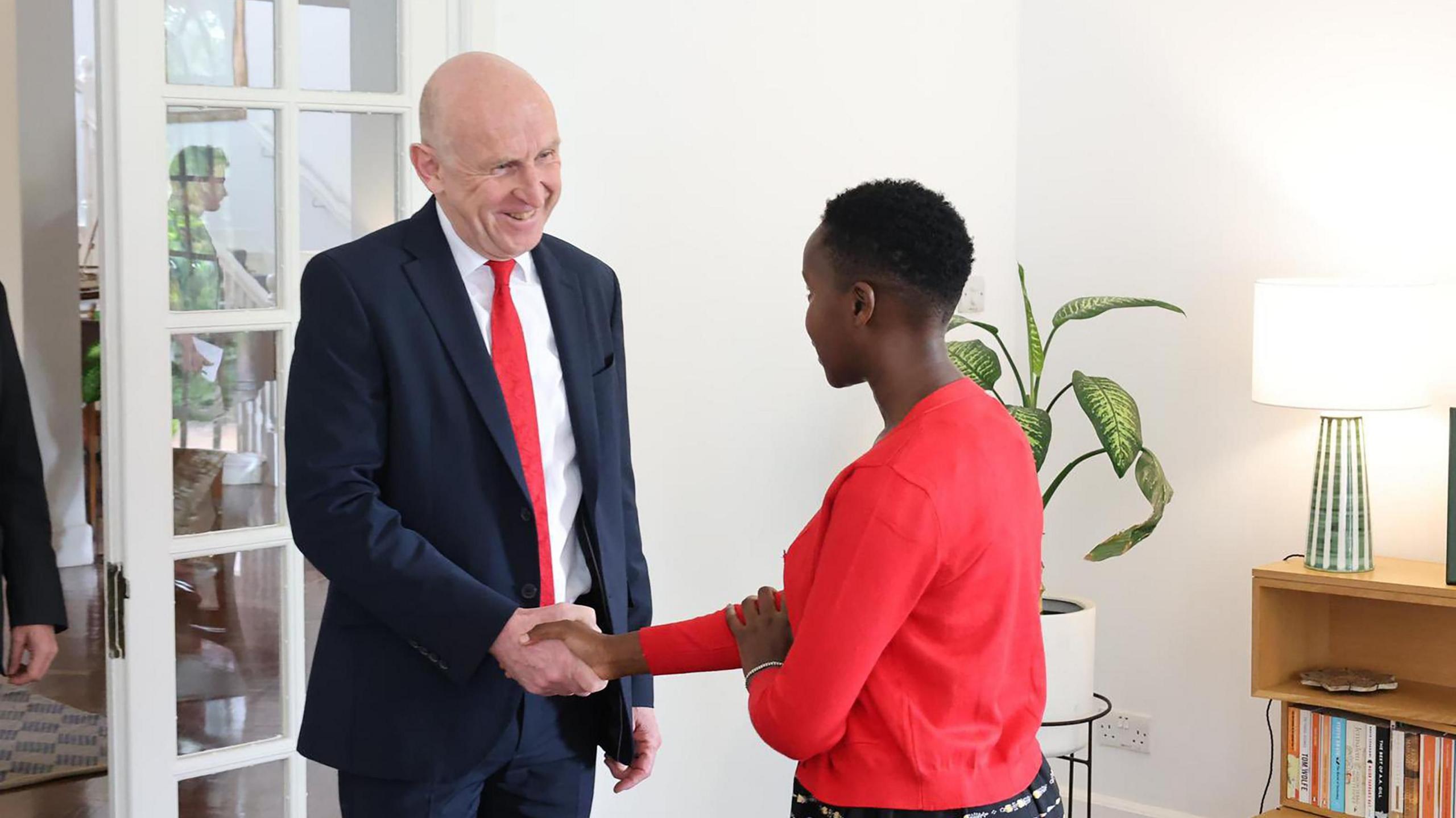 John Healy wearing a dark suit and red tie shaking hands with Esther Njoki.
