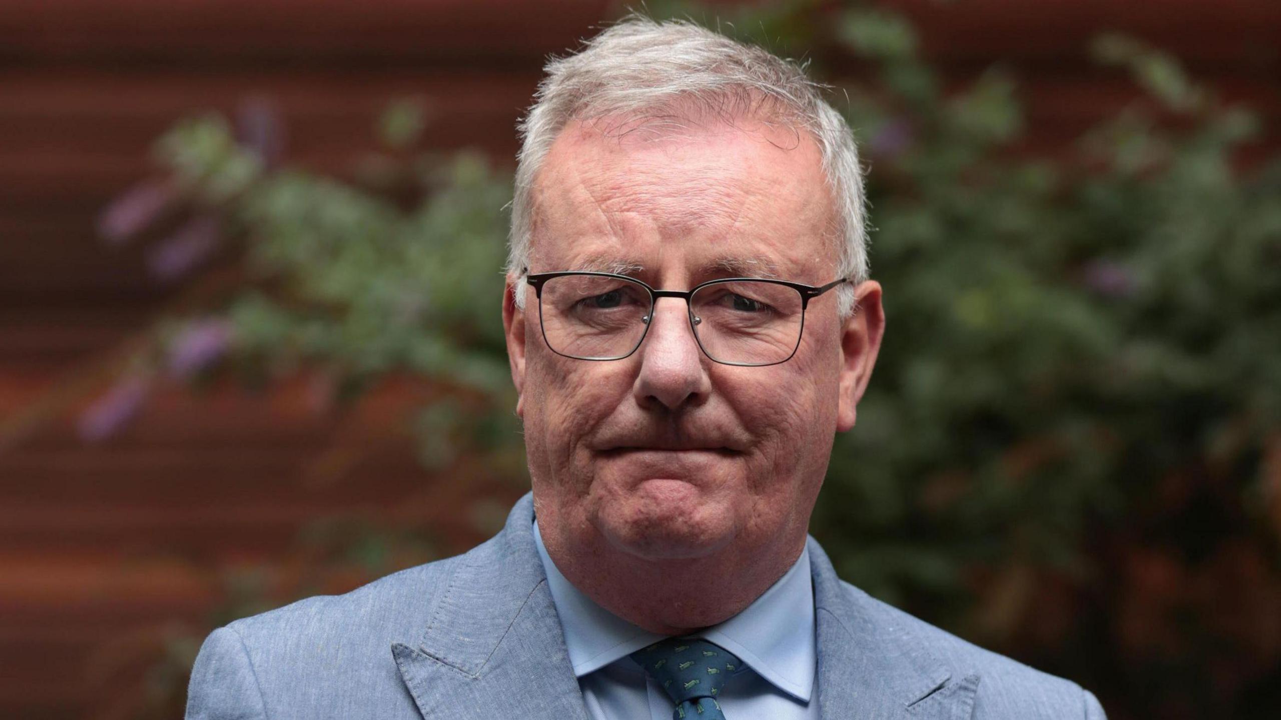 Mike Nesbitt, a man with short, white hair and glasses, looking into the camera during a previous press conference. He is wearing a light grey suit, a light blue shirt and a green patterned tie.