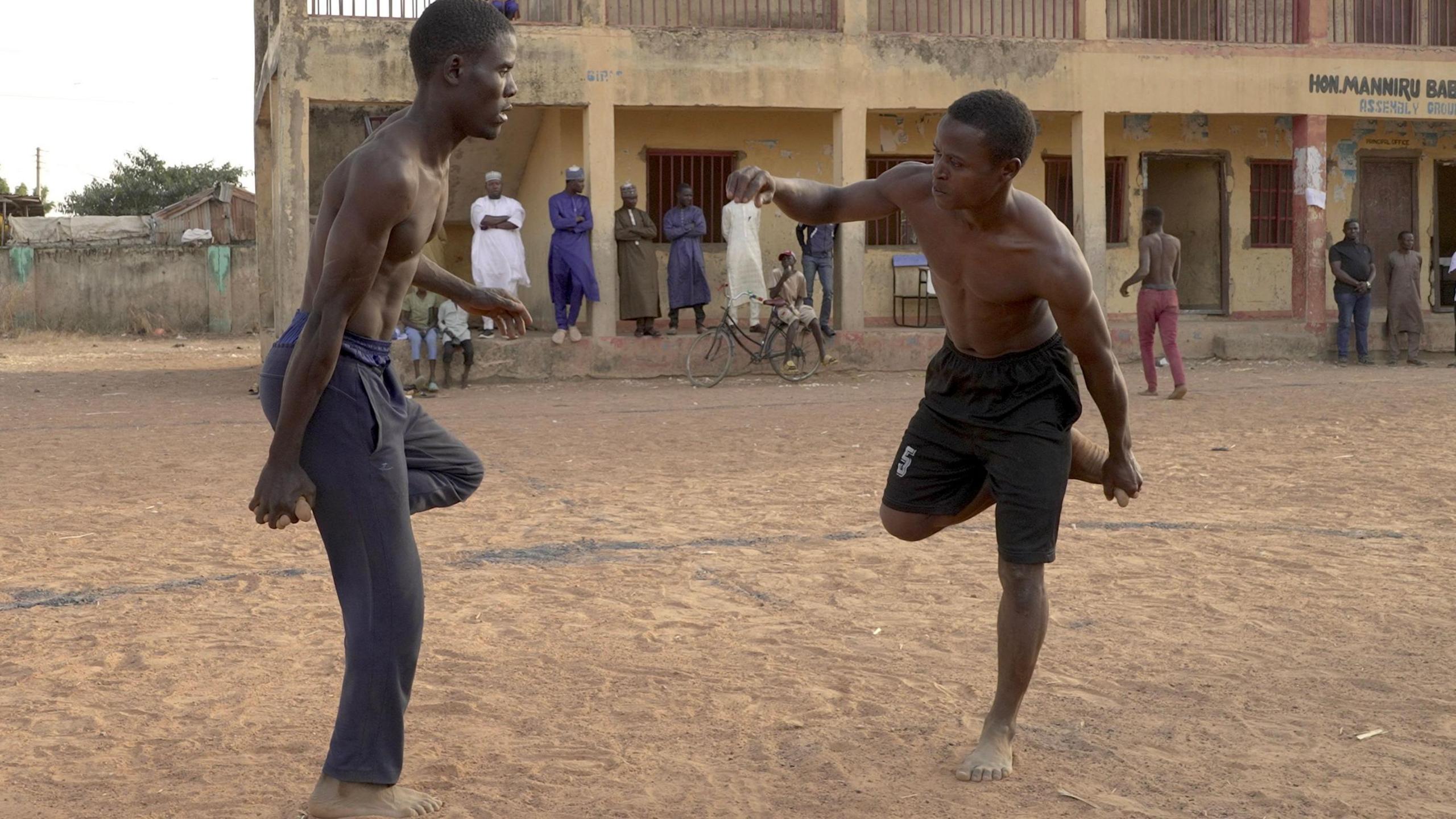 Two topless men are seen opposite each other while balancing on one foot on a dusty patch of land, with several onlookers standing or sitting in front of a two-storey building in the distance