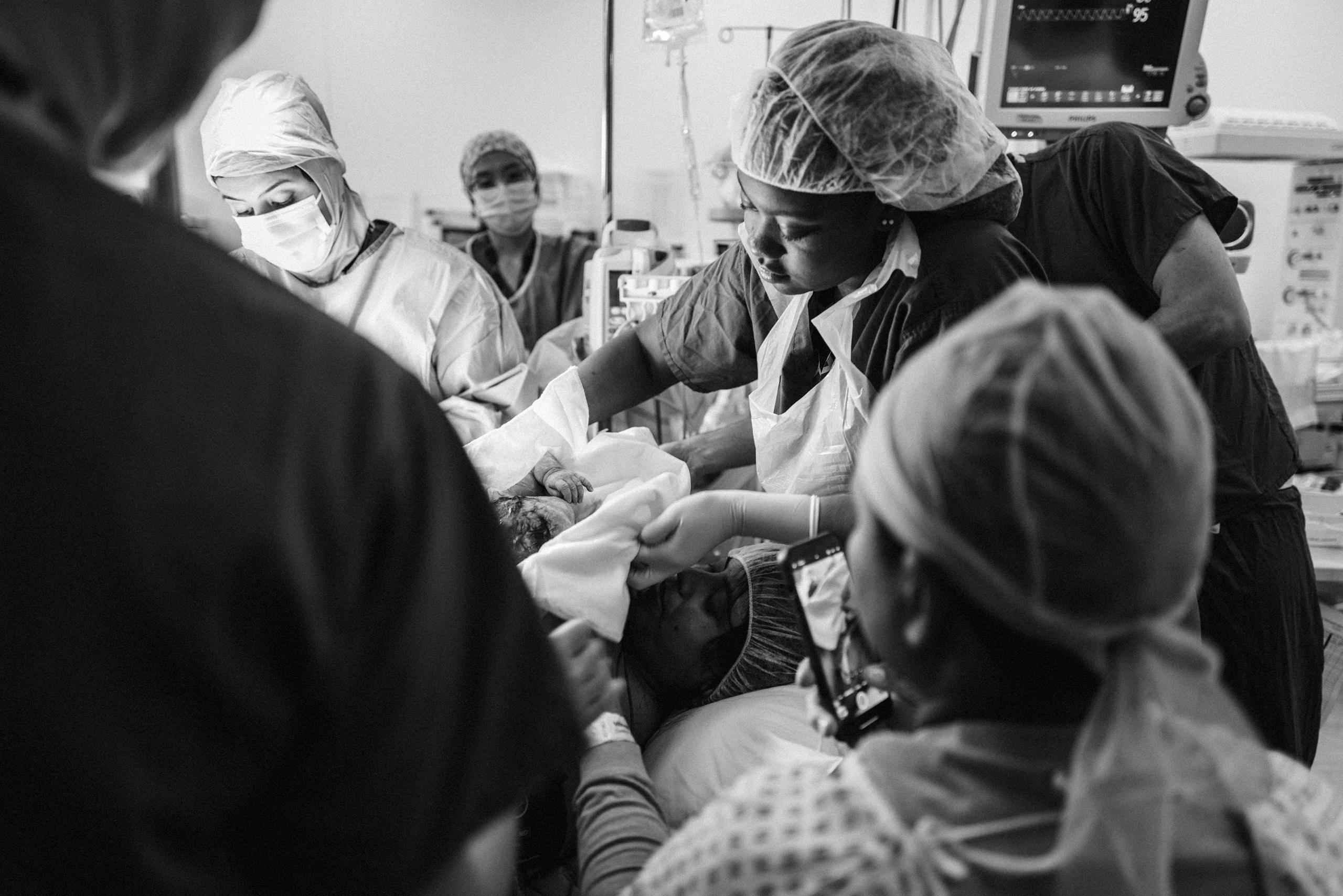 A newborn being placed on the mother’s chest after a C-section, surrounded by medical staff in scrubs and masks. A person captures the moment on their phone.