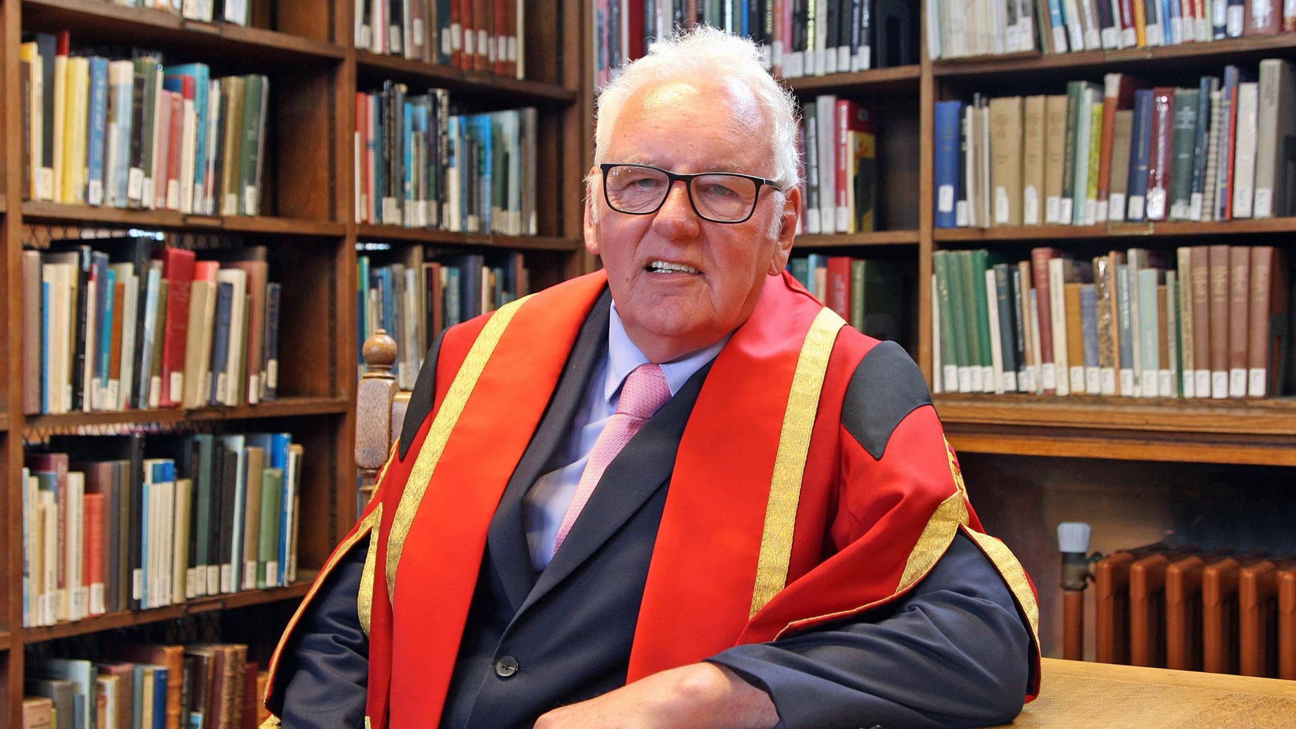 Noel Thomas in his graduation gown, sitting at a wooden table in front of bookshelves