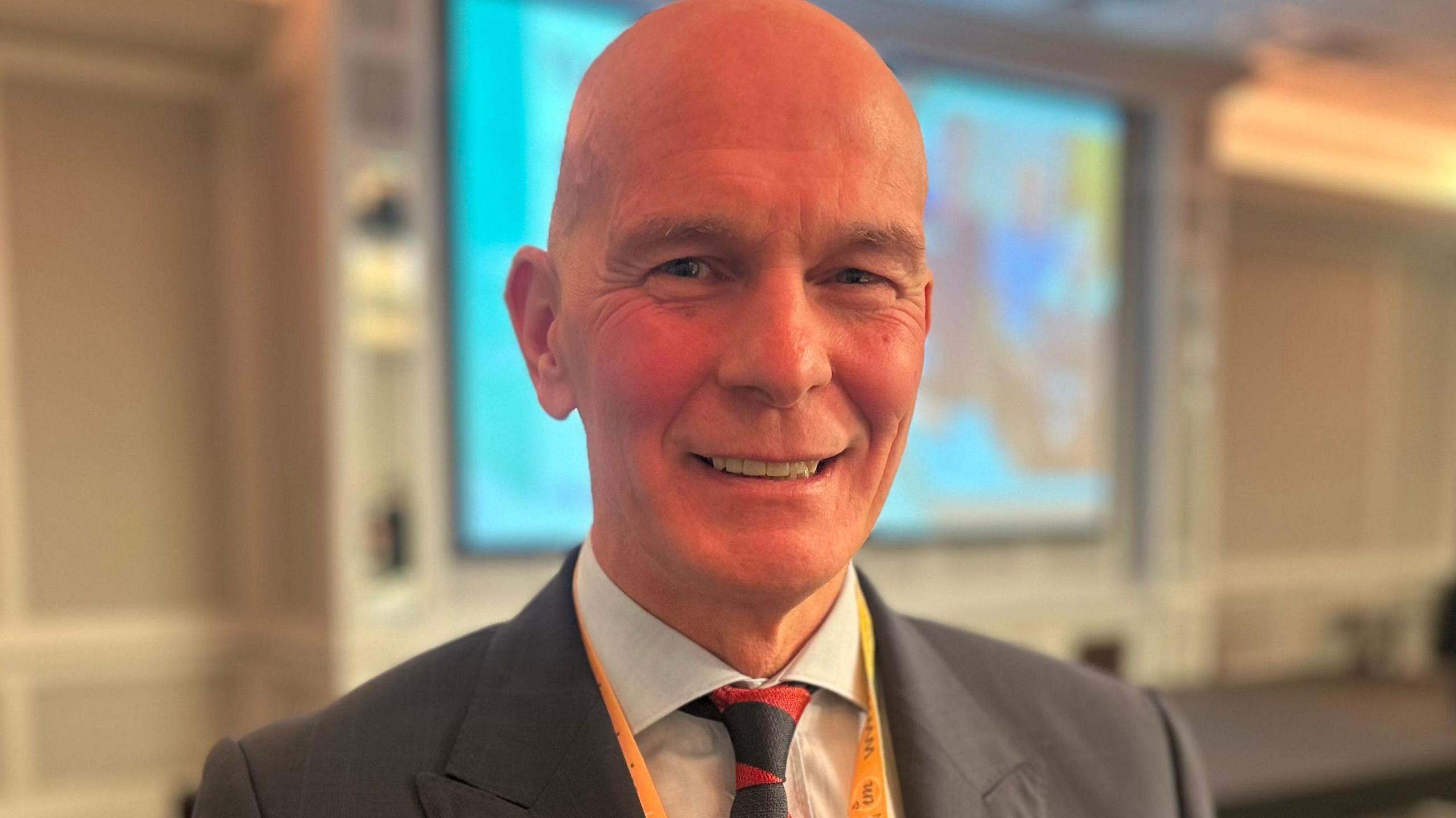 Mark Cox smiles at the conference, he is bald and wears a dark suit jacket, white shirt and red and black tie.