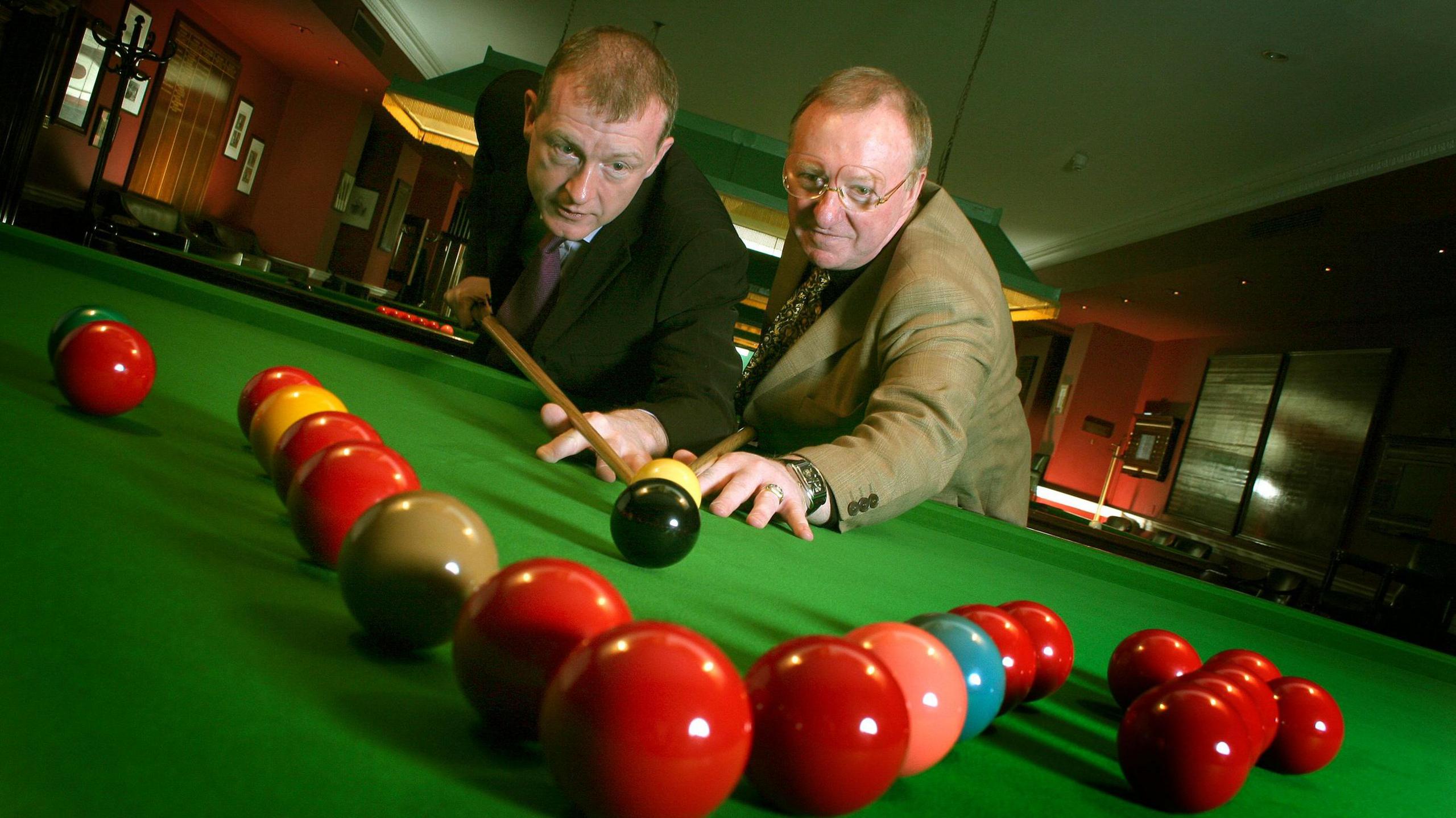 Steve Davis and Dennis Taylor pose at a snooker table, both appear to be aiming for the black ball. There are a number of snooker balls arranged in the front of the image.