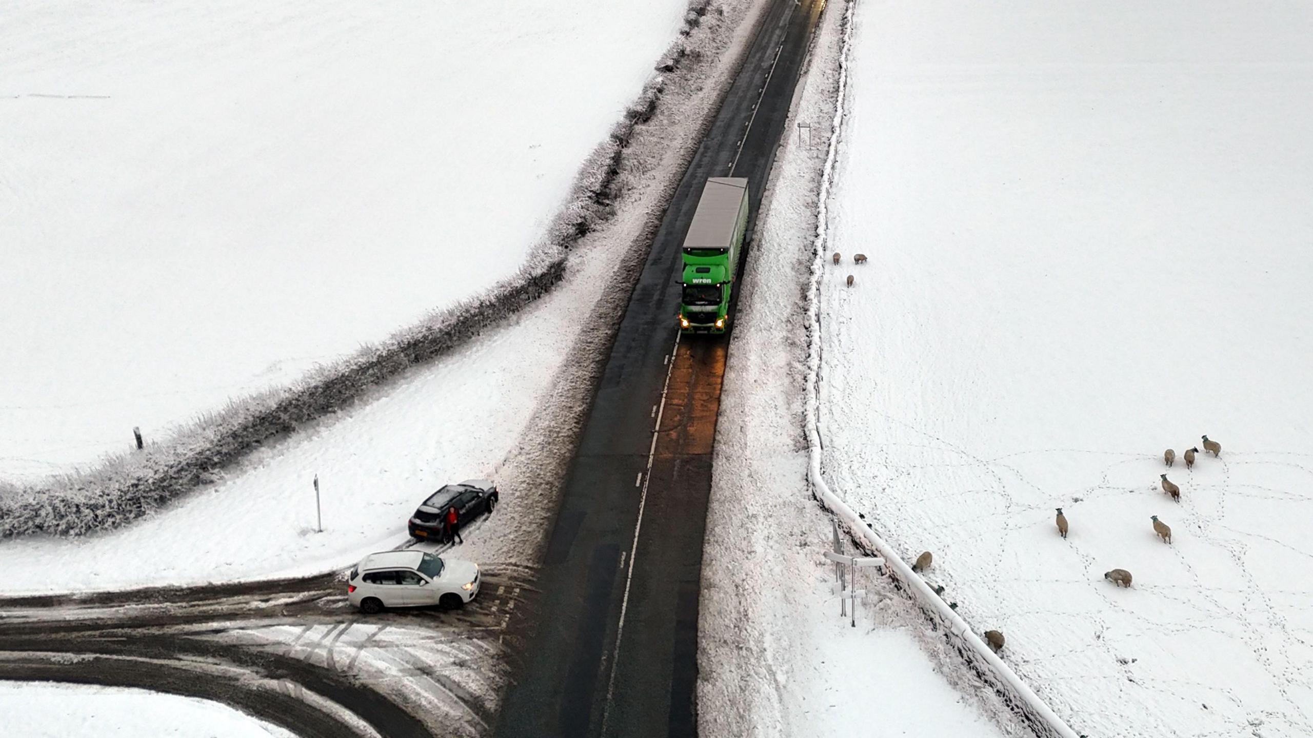 An aerial picture of a road with a truck and several cars driving on it. An intersecting roads has cars waiting to turn onto the road on it. The whole area, apart from the cleared roads, is covered in snow. Sheep are seen in a nearby snow-covered field.