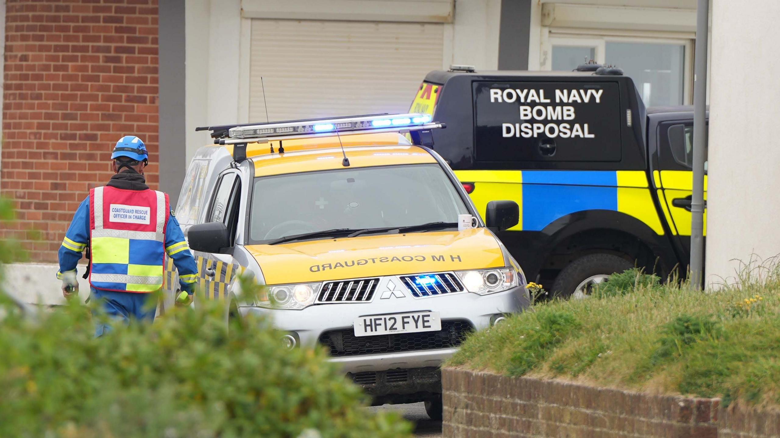 Two four by fours parked close together. One is painted black and has Royal Navy Bomb Disposal on the back, while the other has a yellow bonnet marked HM Coastguard. A man in blue and illuminous yellow safety gear is walking towards the cars.