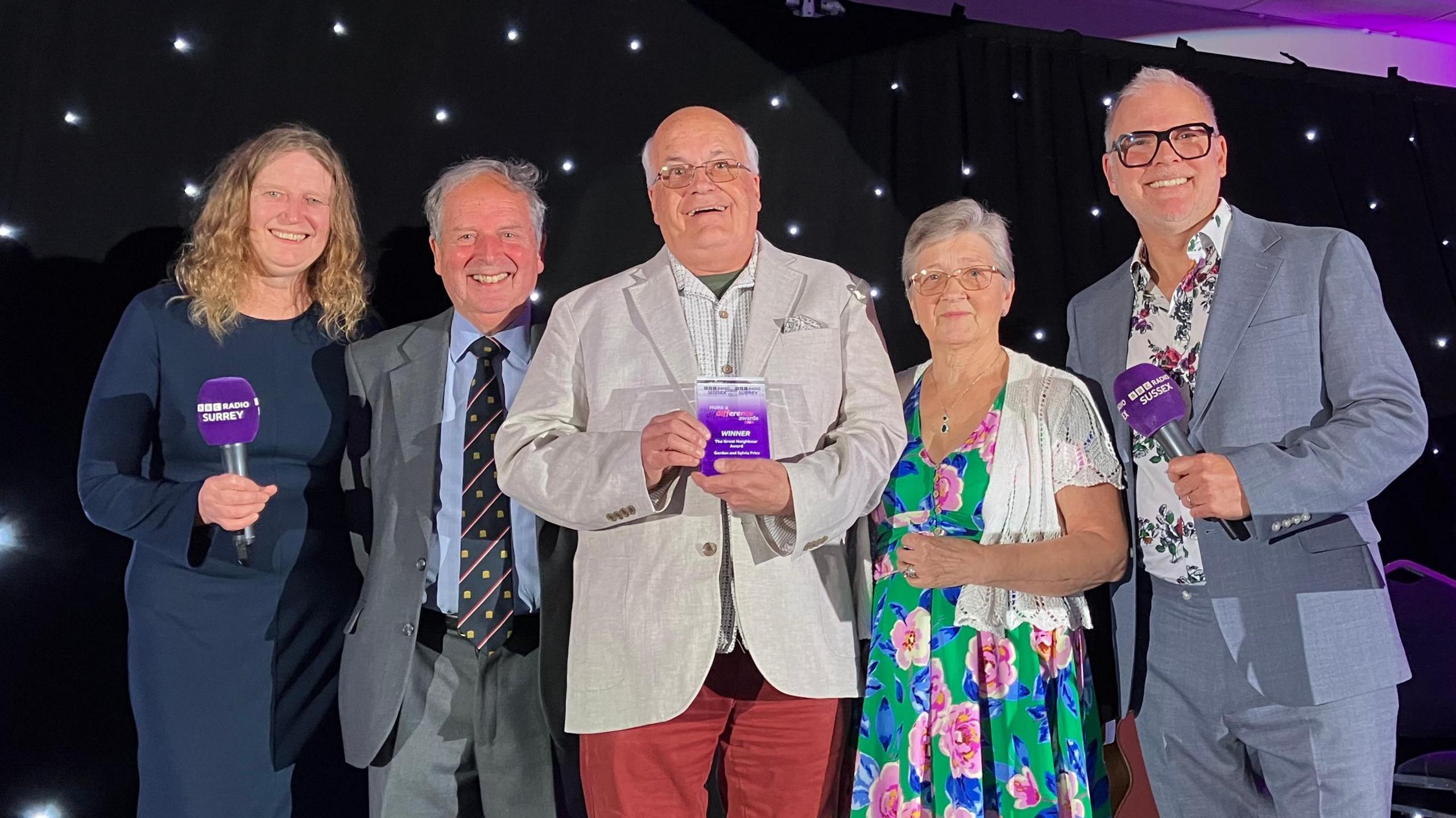 Sarah Gorrell, Canon Peter Bruinvels, Gordon and Sylvia Price and Danny Pike stand on a stage. They are all looking at the camera and smiling, and Gordon is holding their award.