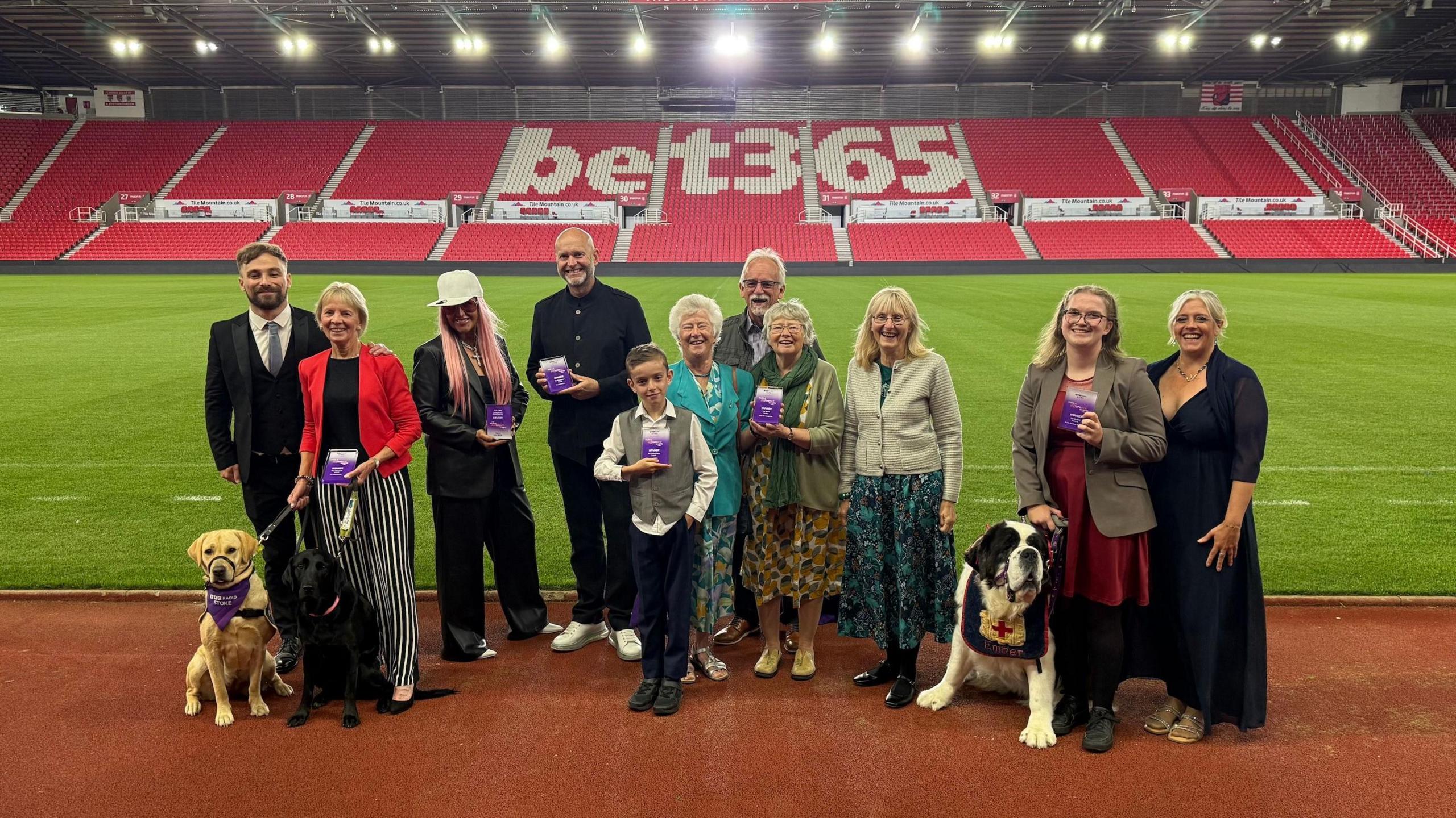 A group of winners from the BBC Radio Stoke Make A Difference Awards are gathered at the side of the pitch at the Bet365 Stadium. There is red branded Bet365 seating behind them and they are stood on the side of the football pitch.