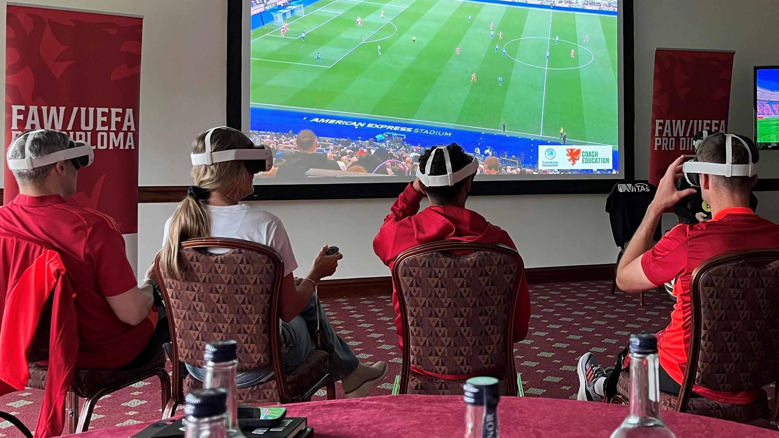 Jonathan Walters (left), Brandi Chastain (second left), Gael Clichy (second right) and Andy King sit wearing virtual reality headsets in front of a large screen.