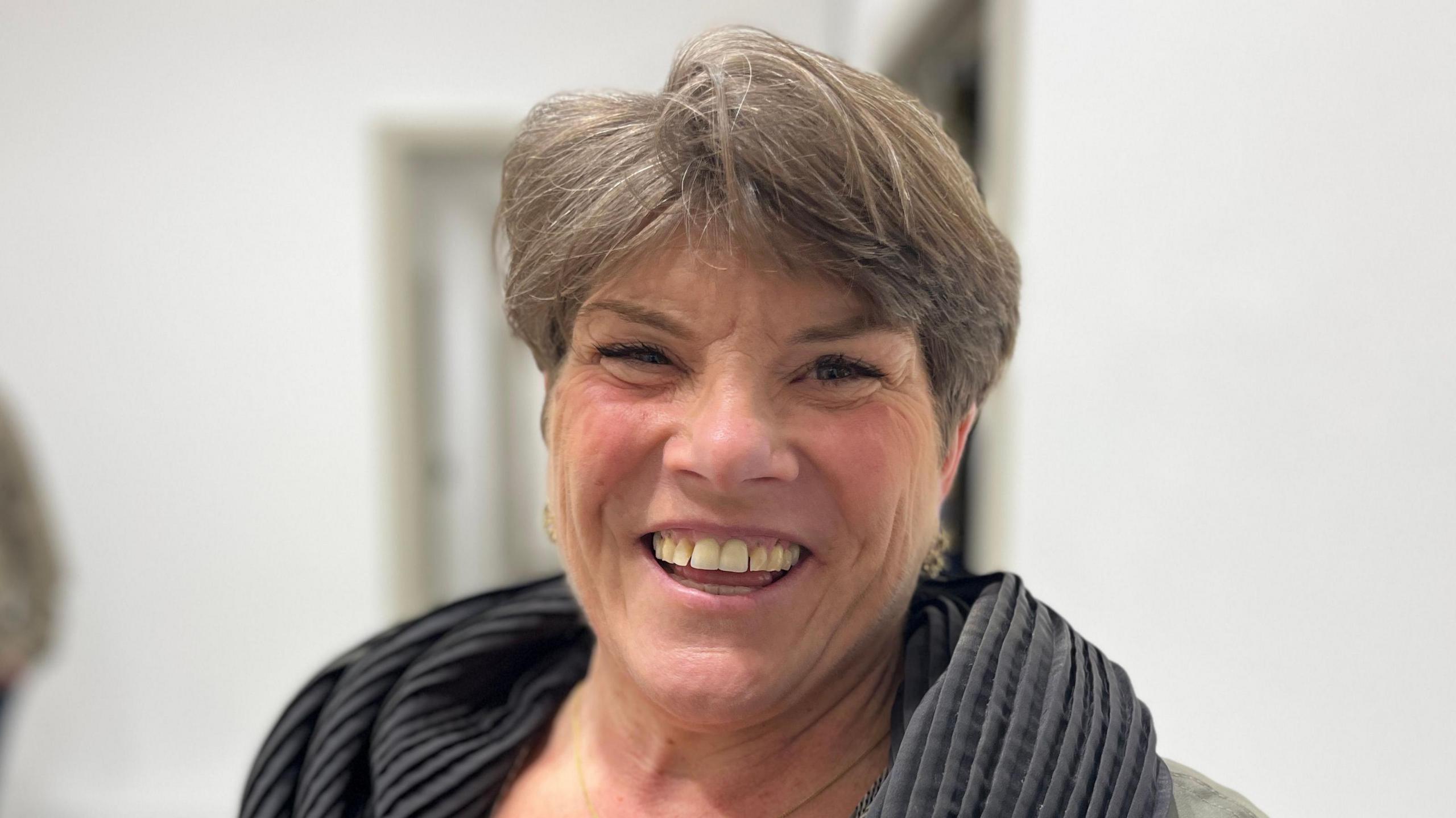 Anna Forgaard has short hair and is wearing a scarf. She is smiling and showing her teeth. She is in focus and the background is blurred. 