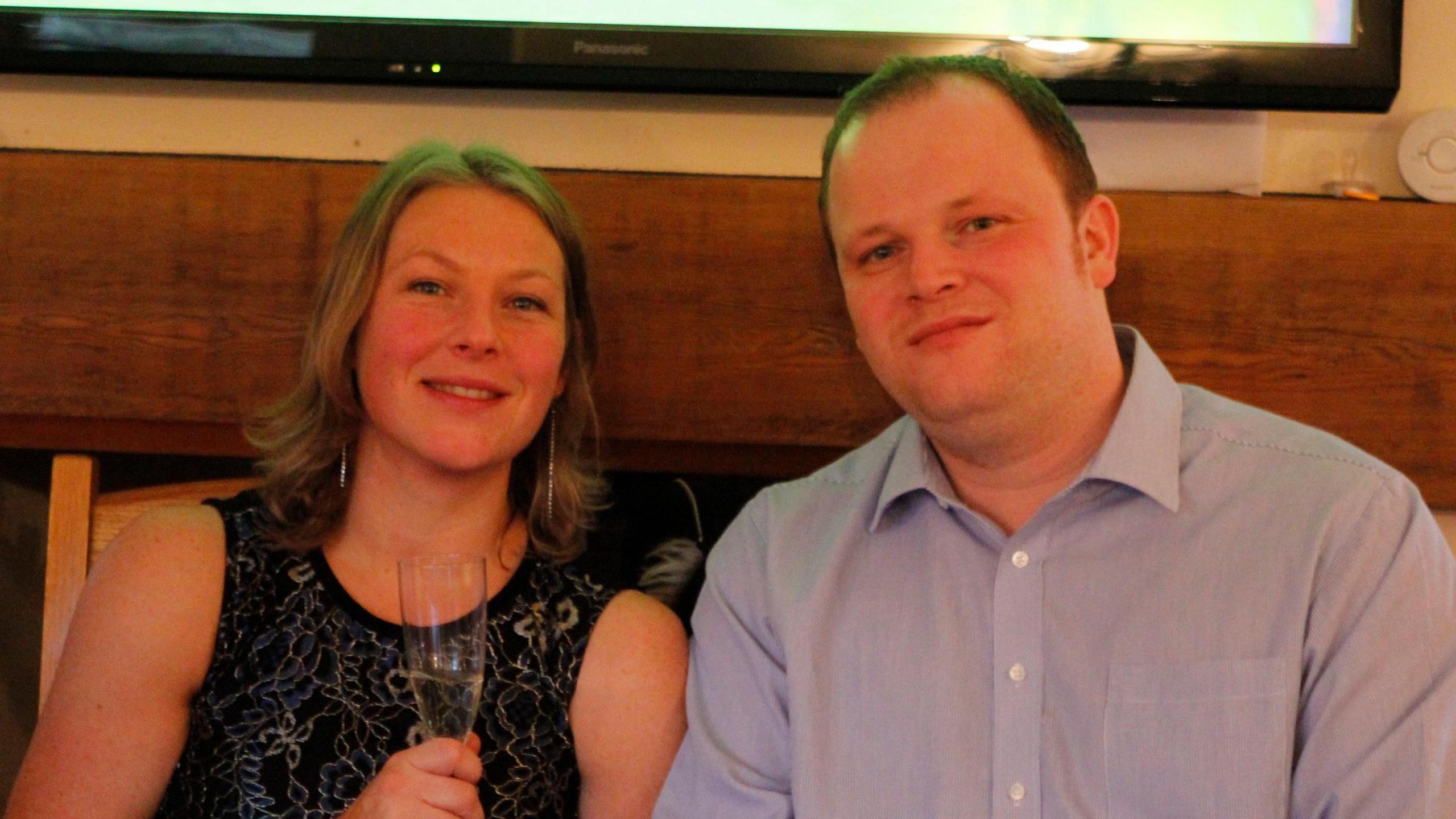 Alison and Rob smile and look at the camera. They are at a party and Alison is drinking from a champagne flute.