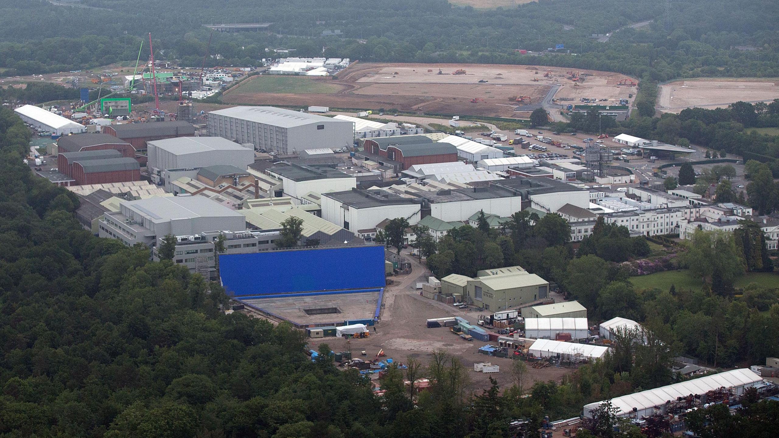 An aerial view of Pinewood Studios. We can see several large grey buildings, with forestry in the foreground.