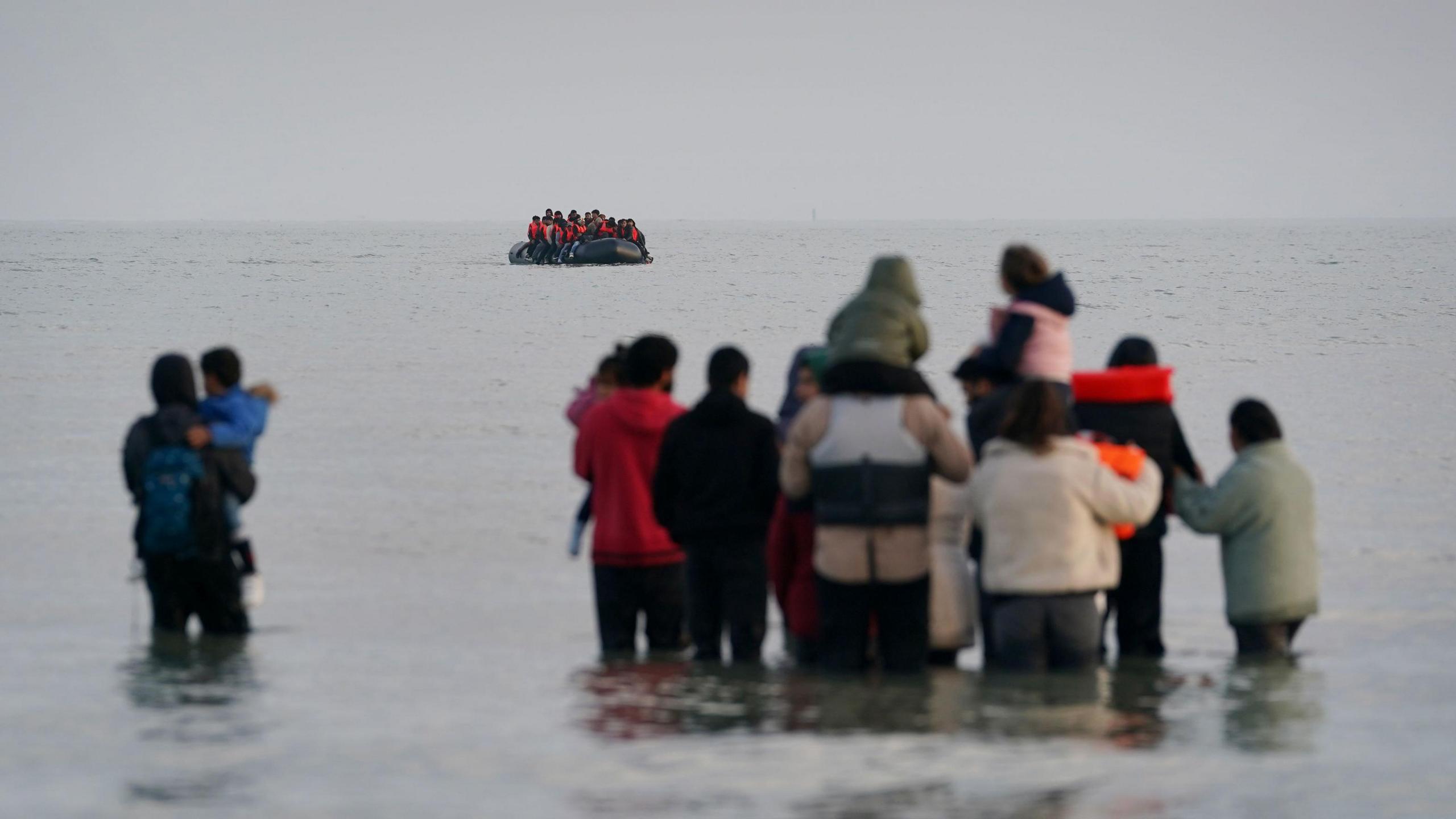 Adults and children standing thigh-deep in the sea. Some children are being carried by adults. Further out to sea is a dinghy packed with people wearing life jackets