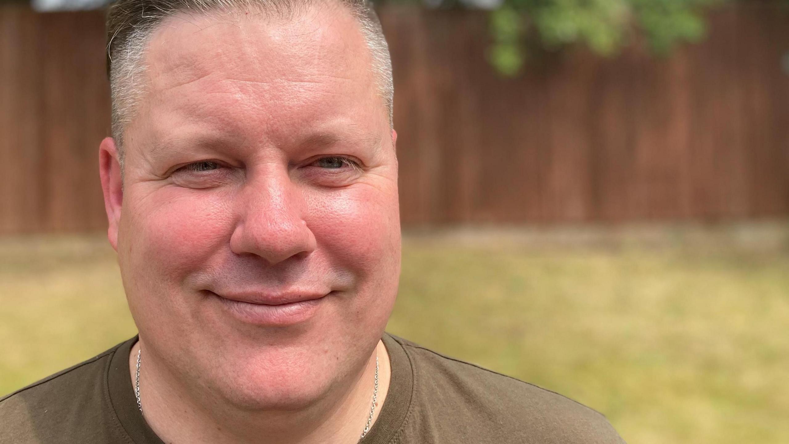 Man smiling and wearing a khaki t-shirt and silver necklace with grass and a fence in the background 