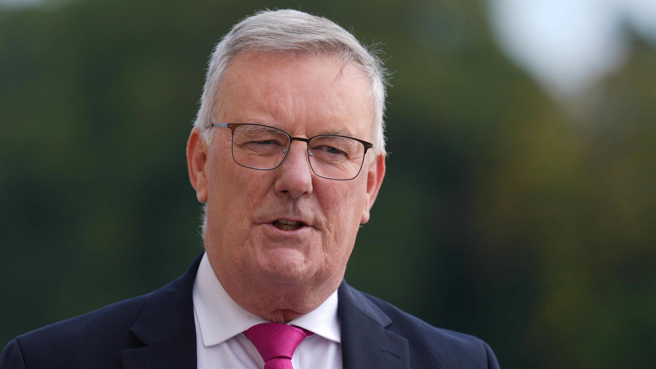 Mike is outside with trees blurred out in the background.
He's wearing glasses, a pink tie, black suit and a white shirt.
He is mid-interview. He has short grey hair. 