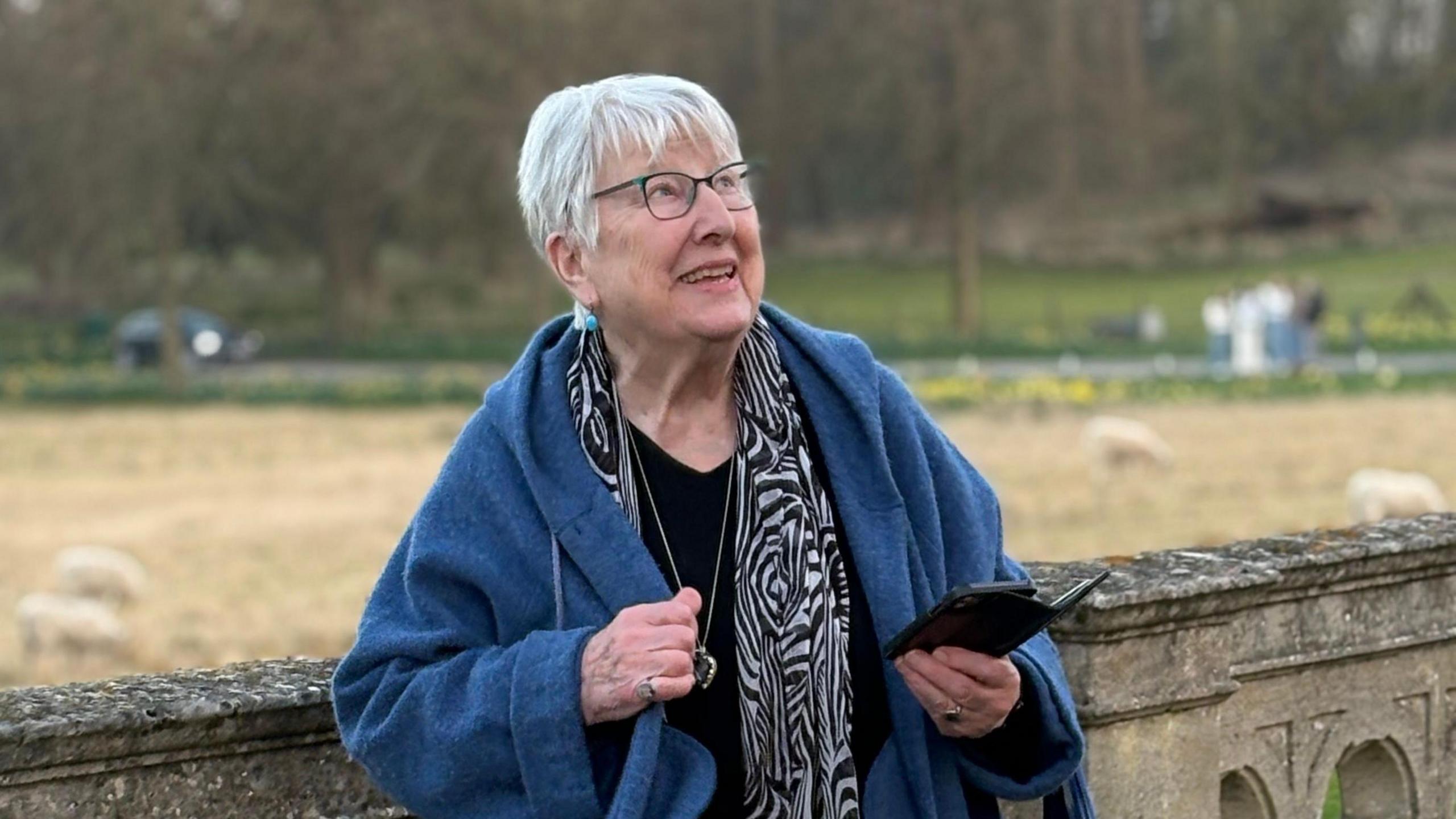 Helen Craig has short, grey hair and is wearing spectacles, a blue coat and a patterned black and white scarf. She is looking skywards and holding a mobile phone. There is a wall behind her and grass in the background. It looks as though she is in the grounds of a stately home