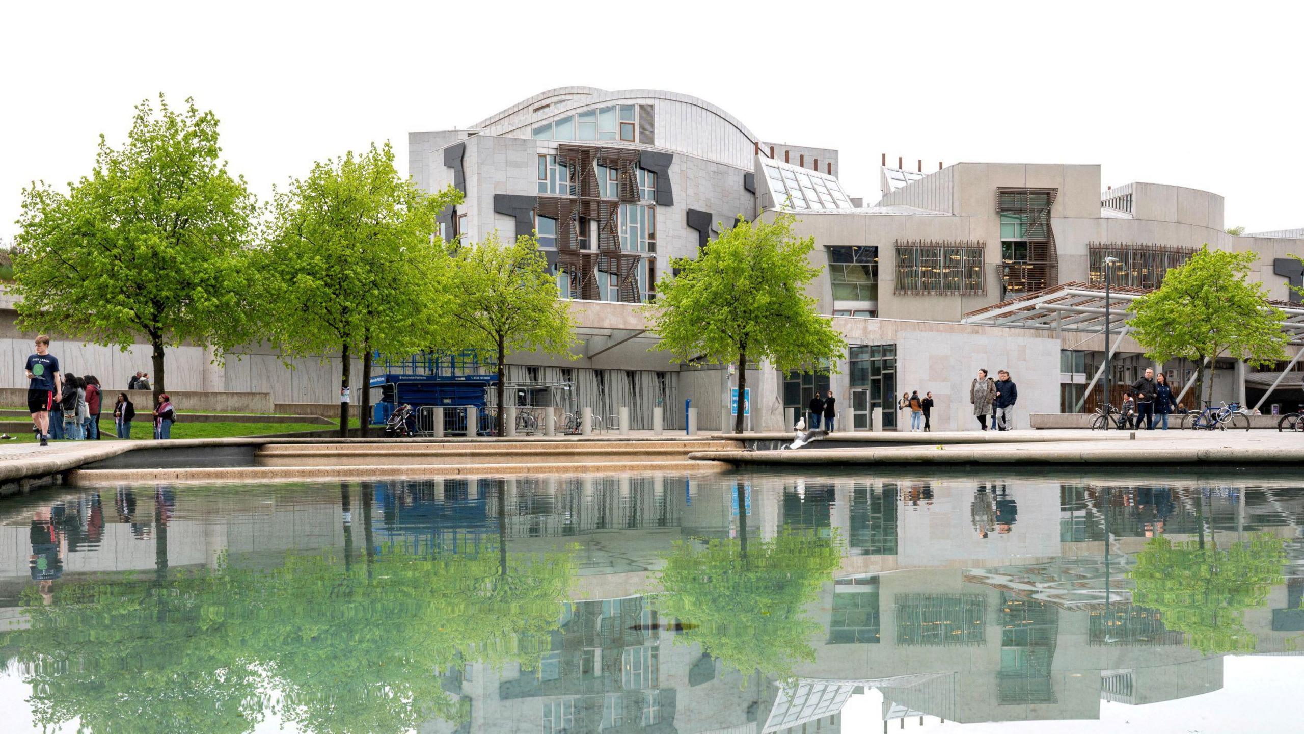 The Scottish Parliament building at Holyrood, with the reflection showing on the water in front of it and people milling by outside