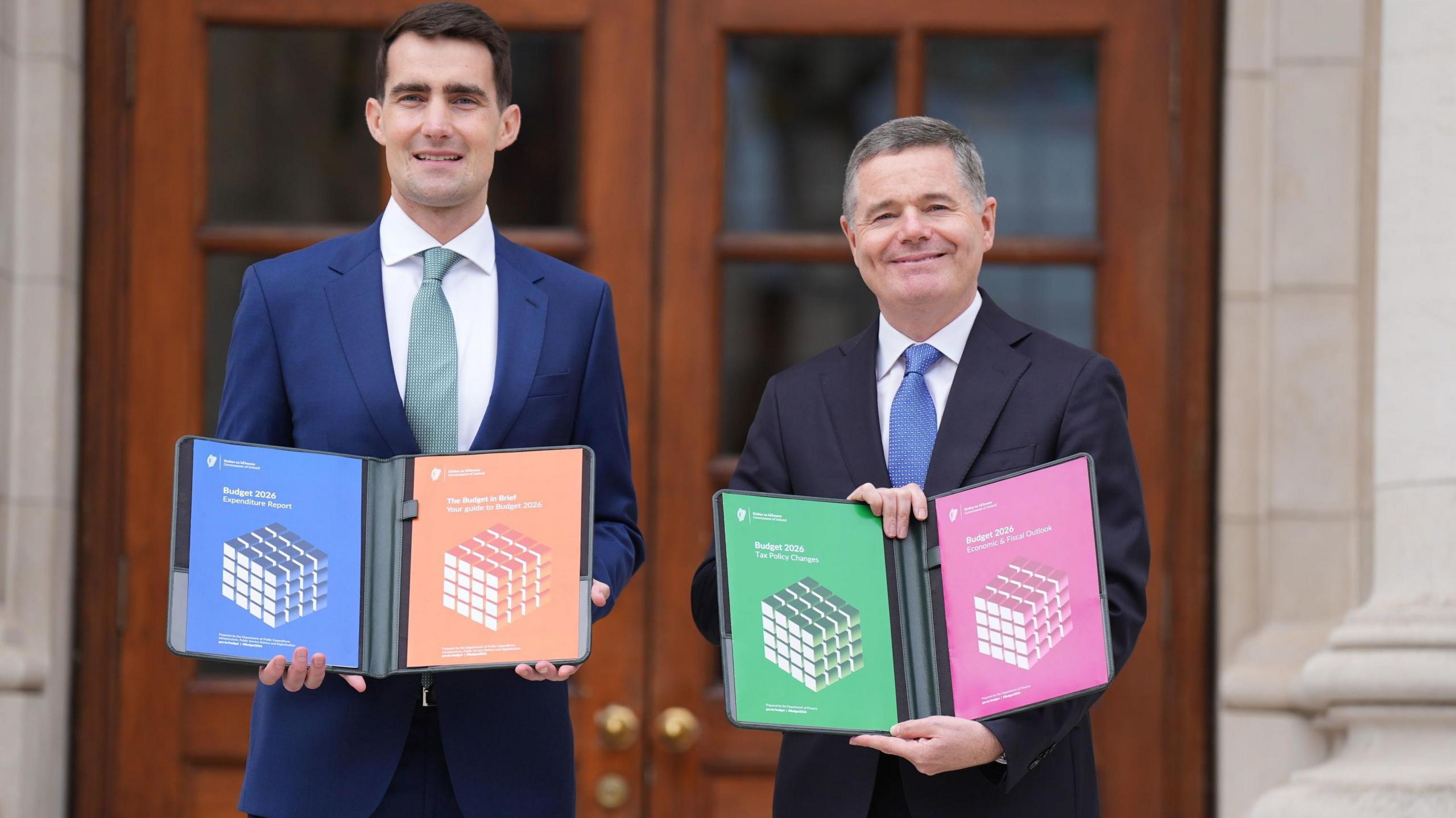 Jack Chambers and Paschal Donohoe hold an open folder containing the main outlines of the budget outside Government building. Both men are in suits and are smiling at the camera.