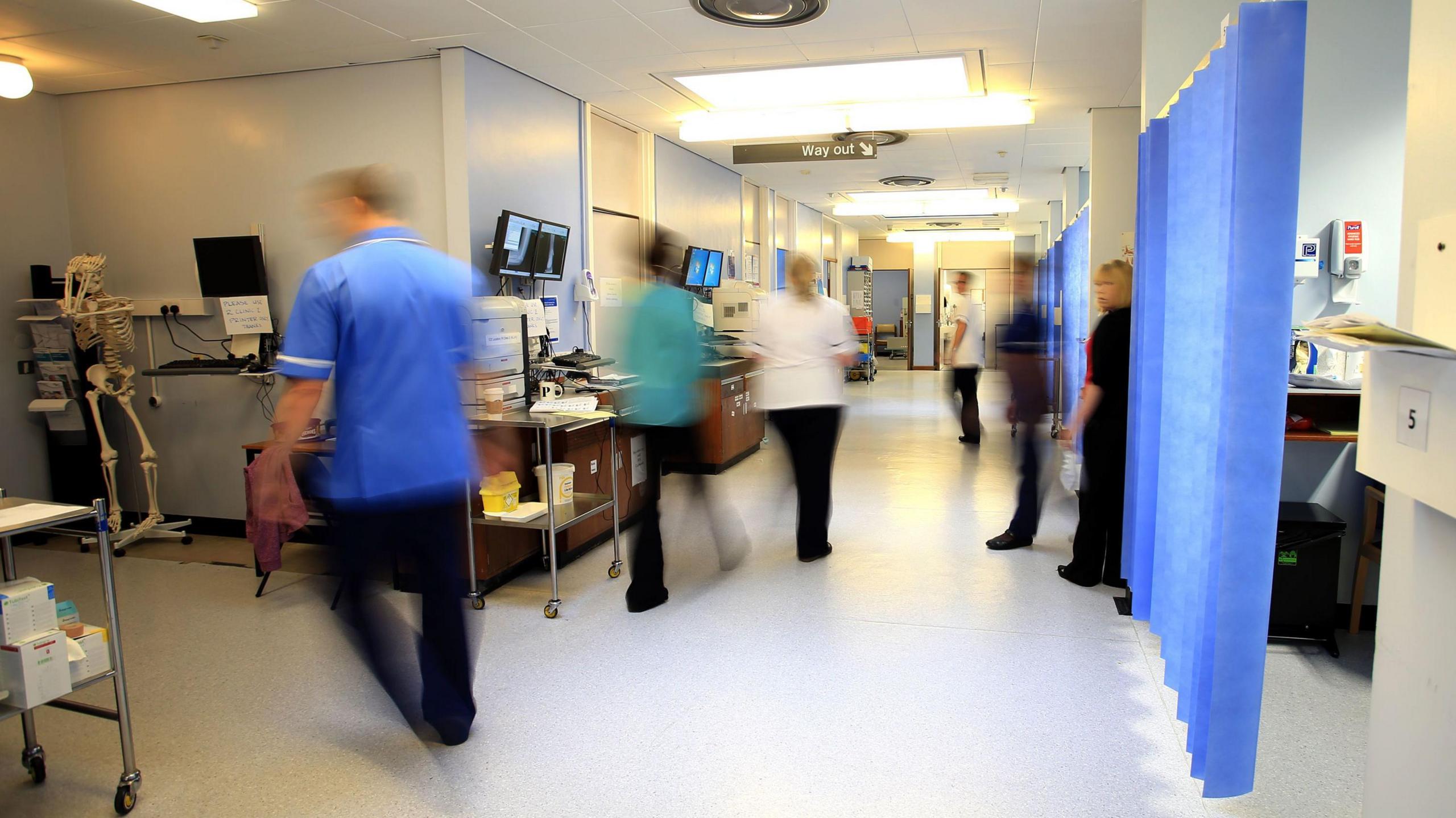 Health care workers wearing blue, green and white scrubs walking around a hospital ward. A set of blue privacy curtains are drawn on the right-hand side. A model skeleton with its hands positioned on its head is on the left.