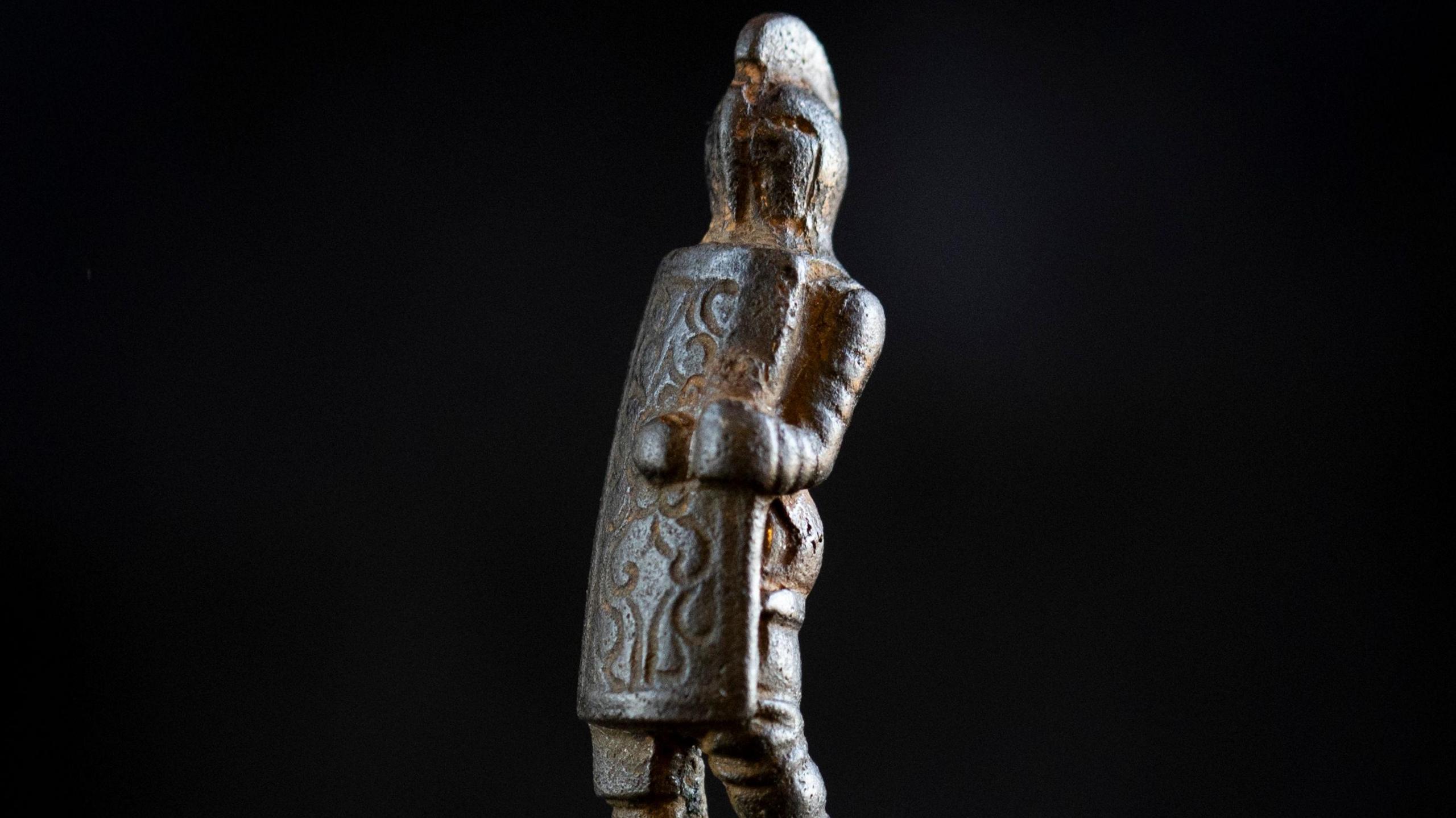 A metal knife handle which is carved into a gladiator shape. The figurine is wearing a helmet and holding a patterned shield which covers most of its body.