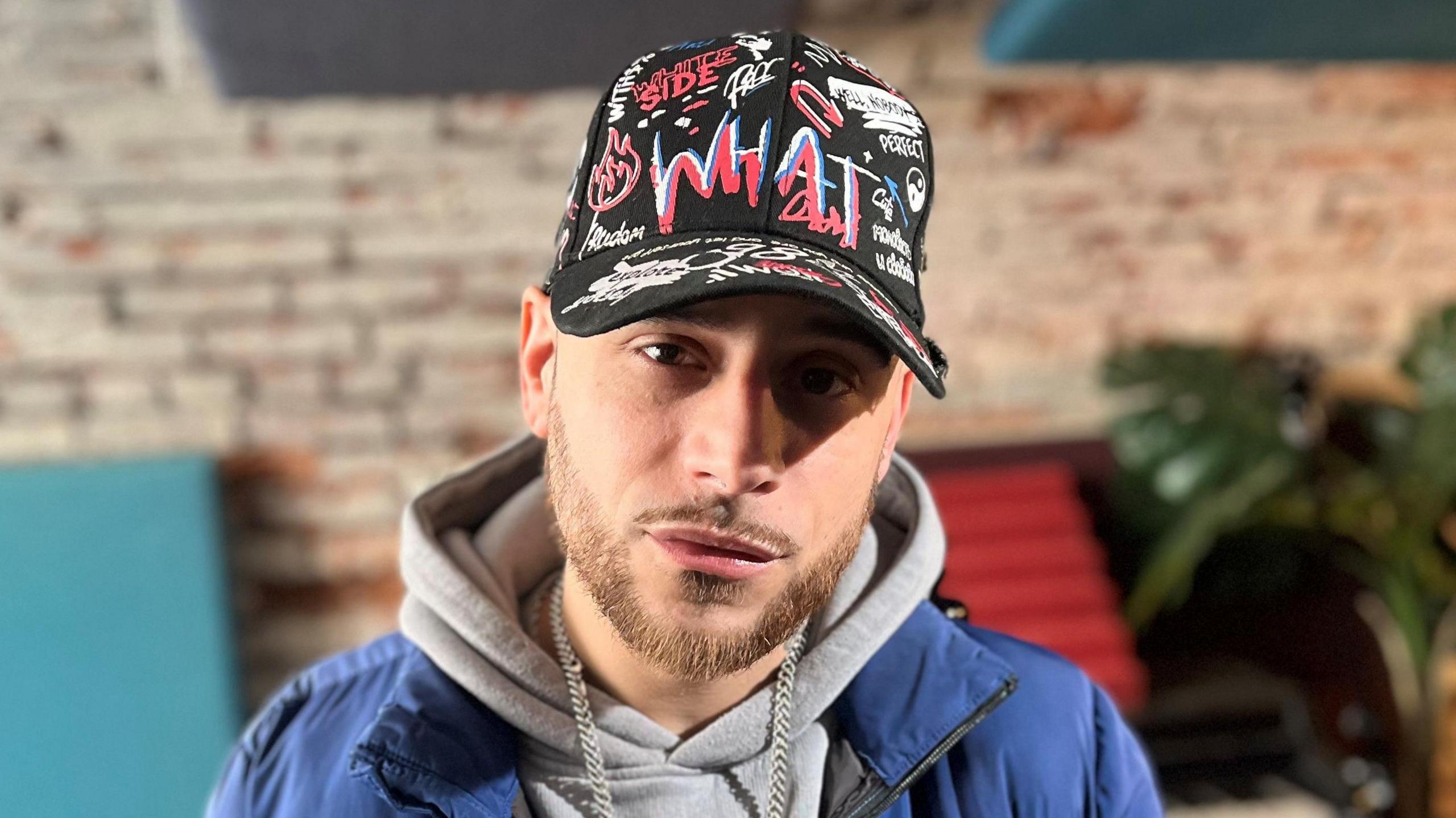 JT, a man in his thirties wearing a blue puffer coat and a grey hoodie. He is wearing a black hat with graffiti printed on it. He has a bit of facial hair. The background is out of focus but a brick wall and a green plant can be seen.